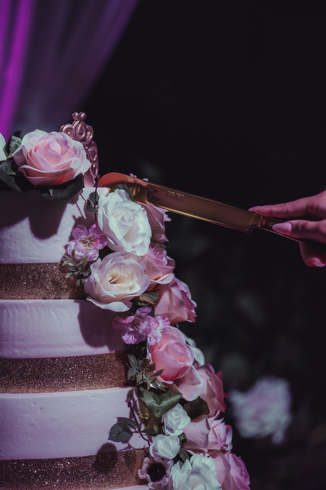 Hand cutting a multi-tiered white wedding cake adorned with pink and white roses in elegant purple lighting.