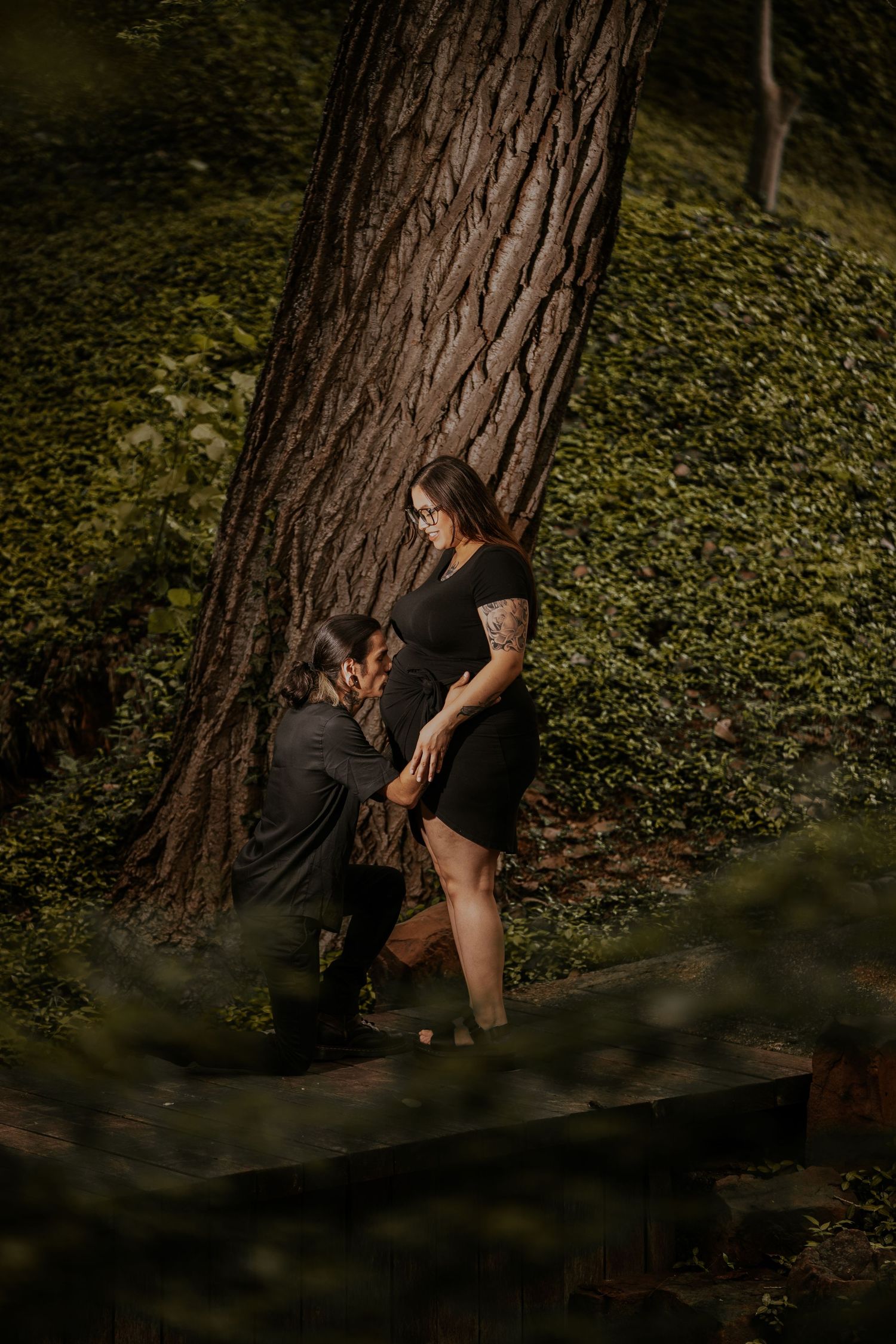 A couple shares an intimate moment against a tree trunk in a wooded forest setting with dappled lighting.