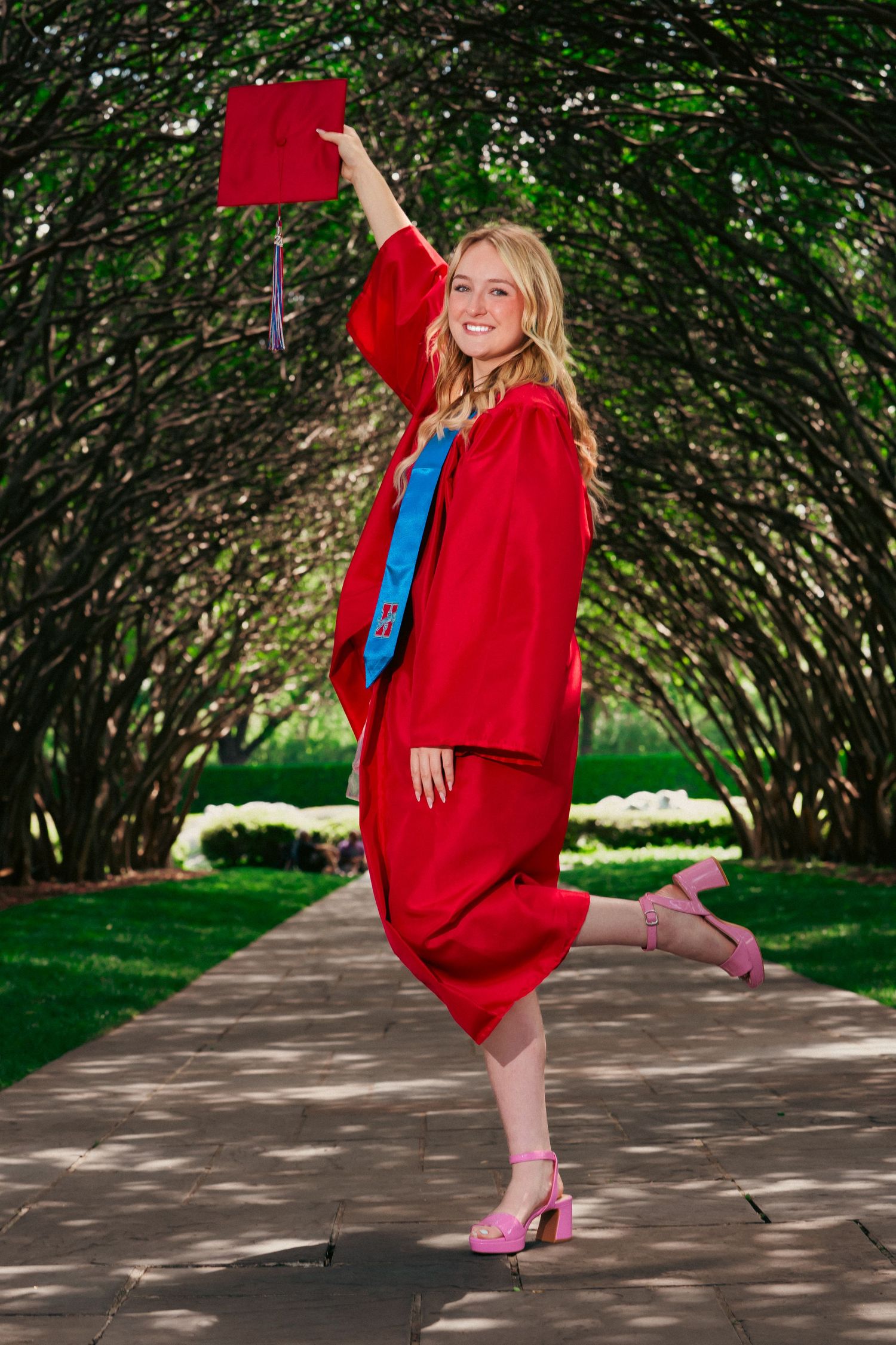 A figure in a vibrant red graduation gown dances and poses playfully on a tree-lined path in a garden setting.