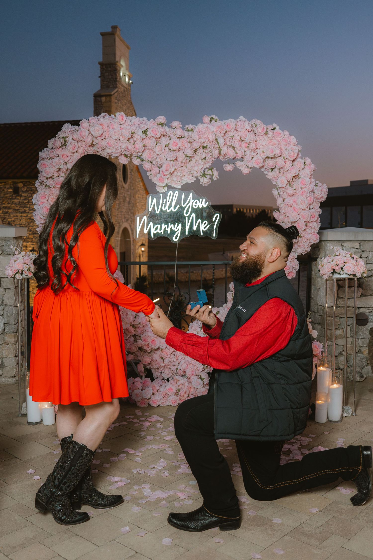 A man proposes on one knee to a woman in a red dress beneath a floral heart arch with a Will You Marry Me sign.