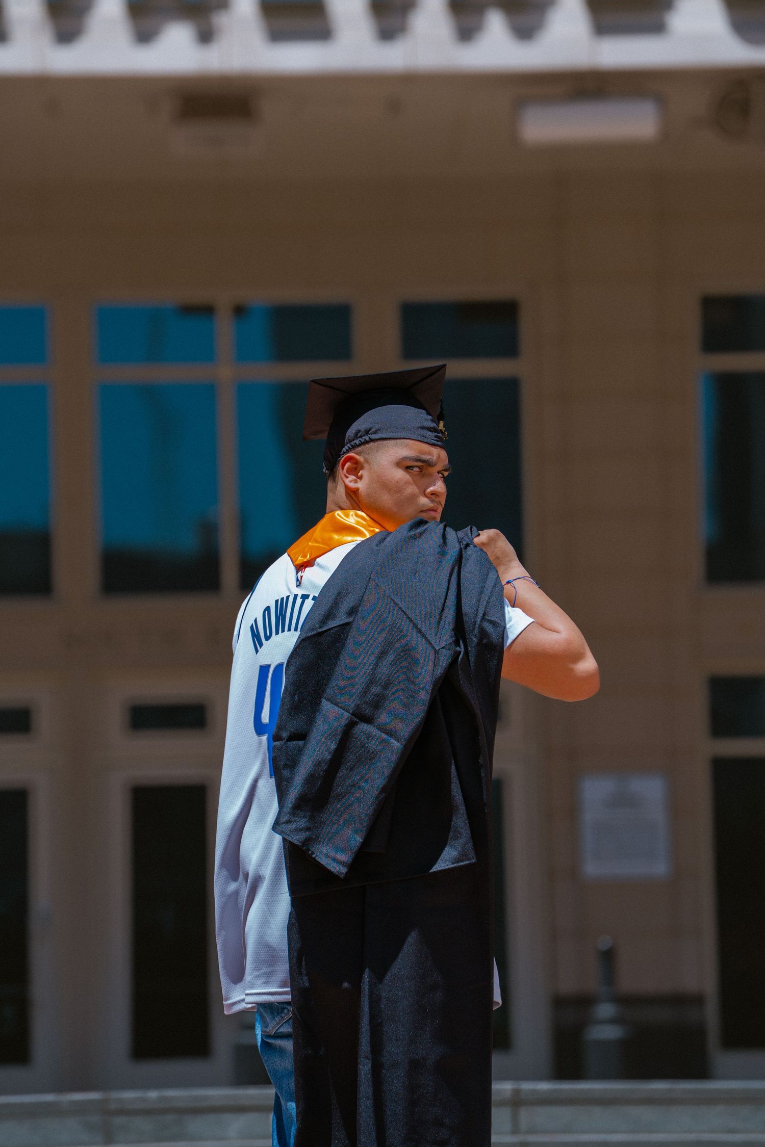 Graduate in cap and gown holding robe over shoulder wearing Nowitzki Dallas jersey outdoors.
