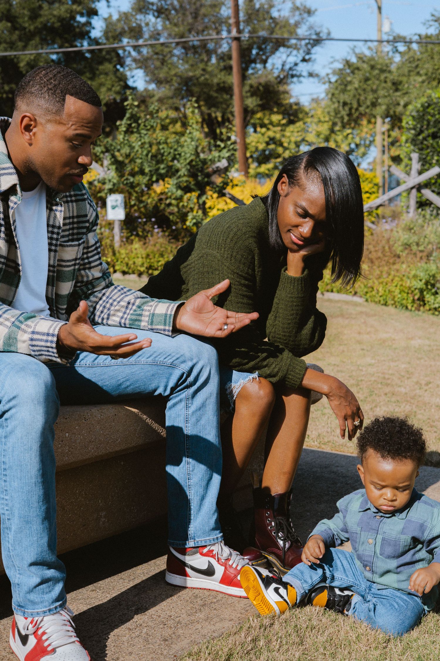 Black parents interacting with toddler sitting on ground in sunny park, mom in green sweater smiling.