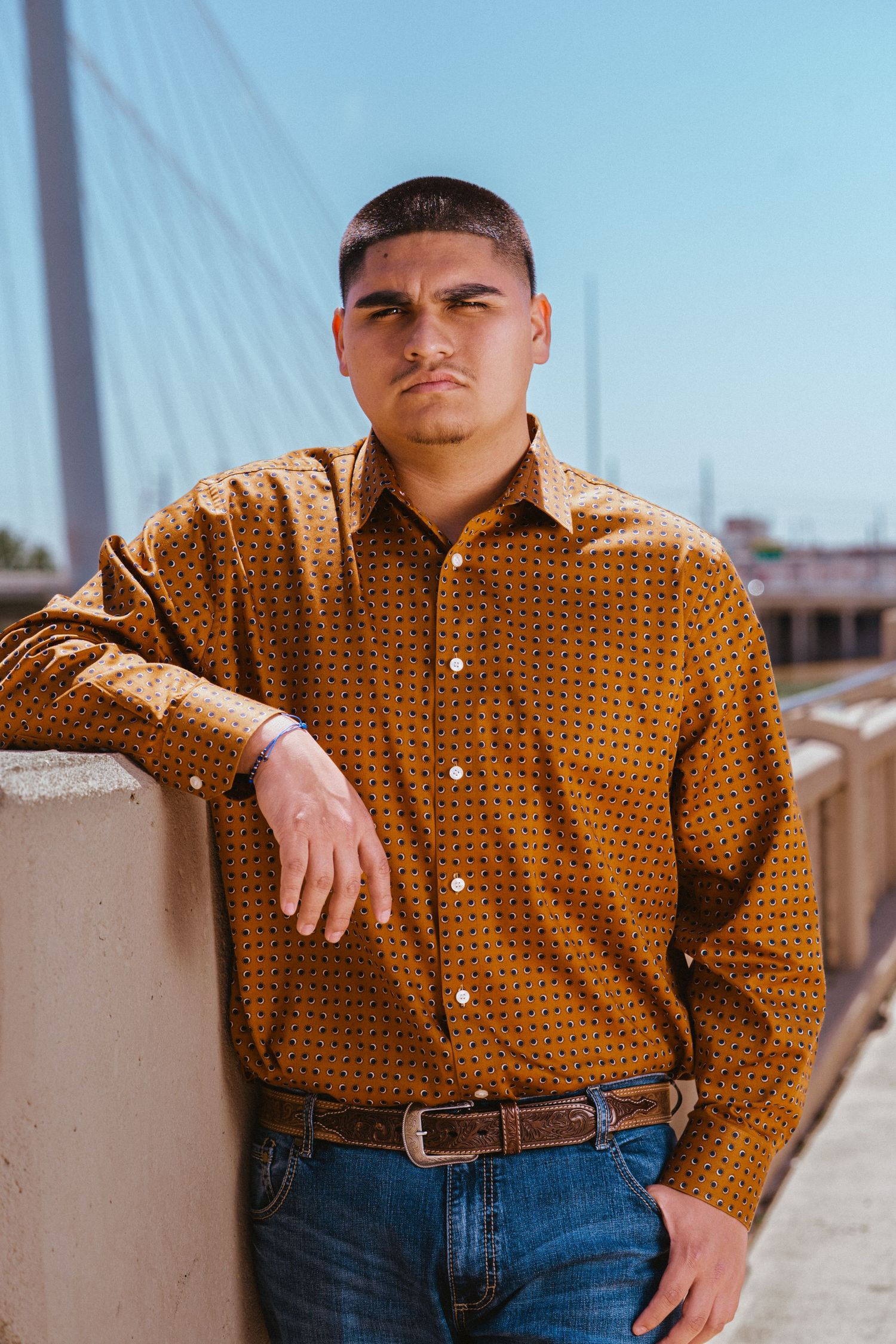 Young man in mustard button-up shirt leaning against wall near suspension bridge on sunny day.