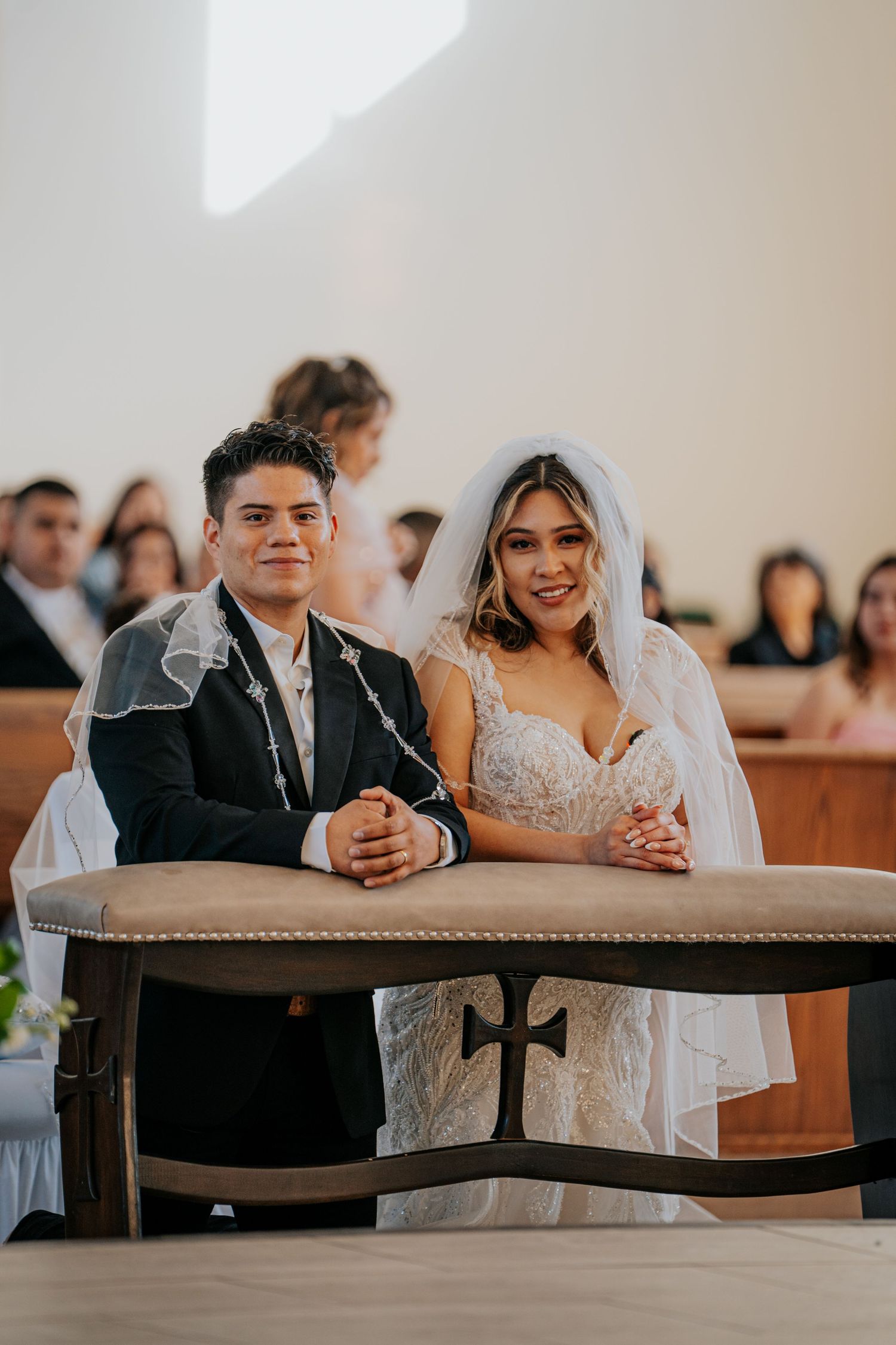 Newlyweds sit together in a church pew during their wedding ceremony, smiling and sharing a joyful moment.
