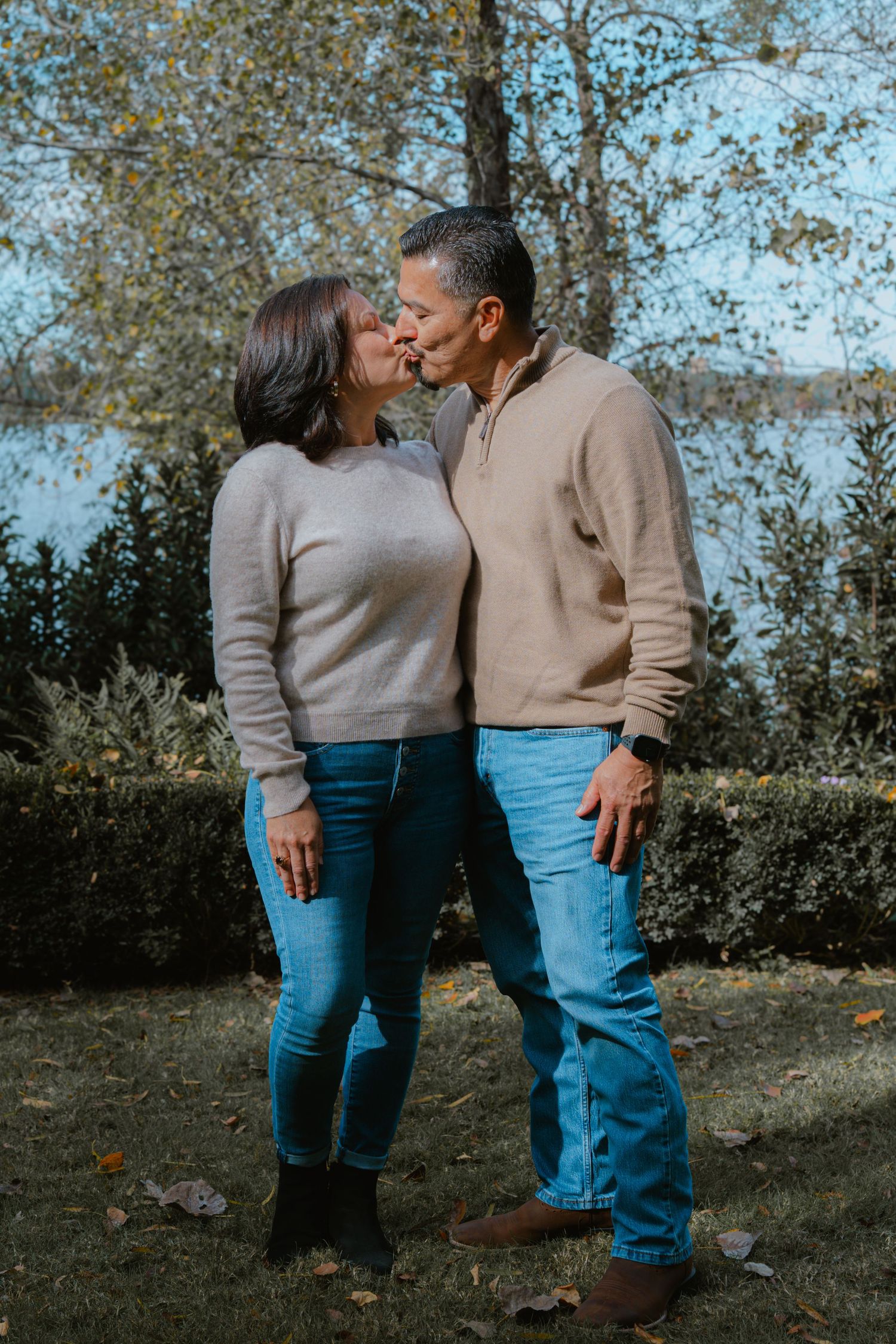 A couple wearing matching beige sweaters and blue jeans share an intimate moment outdoors during autumn.