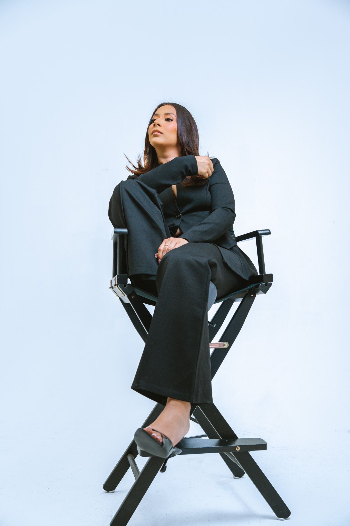 A person in a black outfit poses artistically on a dark director's chair against a white background.
