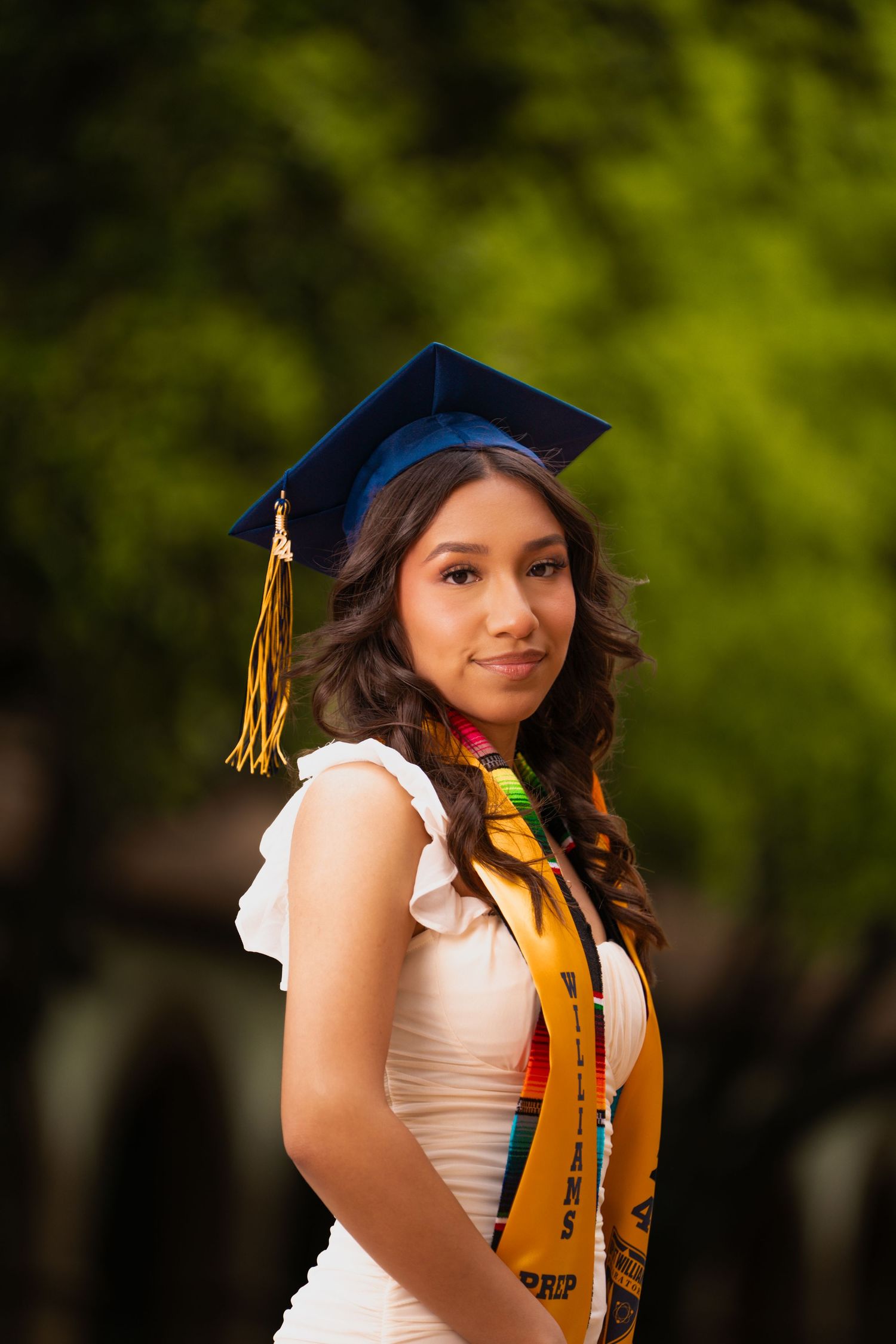 Graduate in white dress and cap with gold tassel poses against blurred green background during graduation photoshoot.