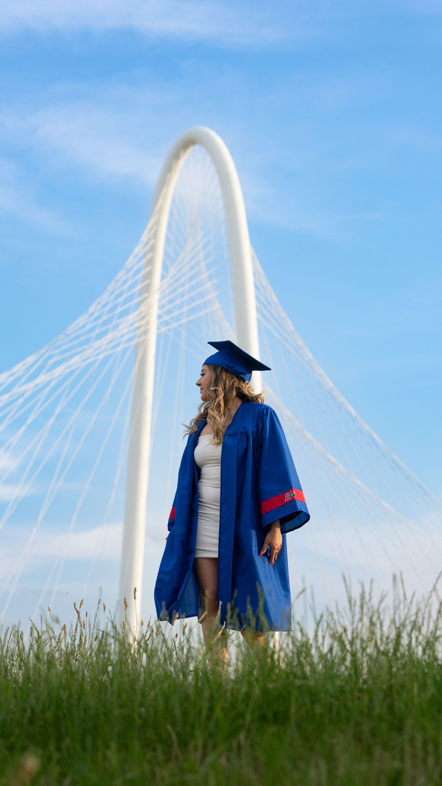 A figure in a blue graduation gown stands against a blue sky backdrop with a modern white bridge arch in the distance.