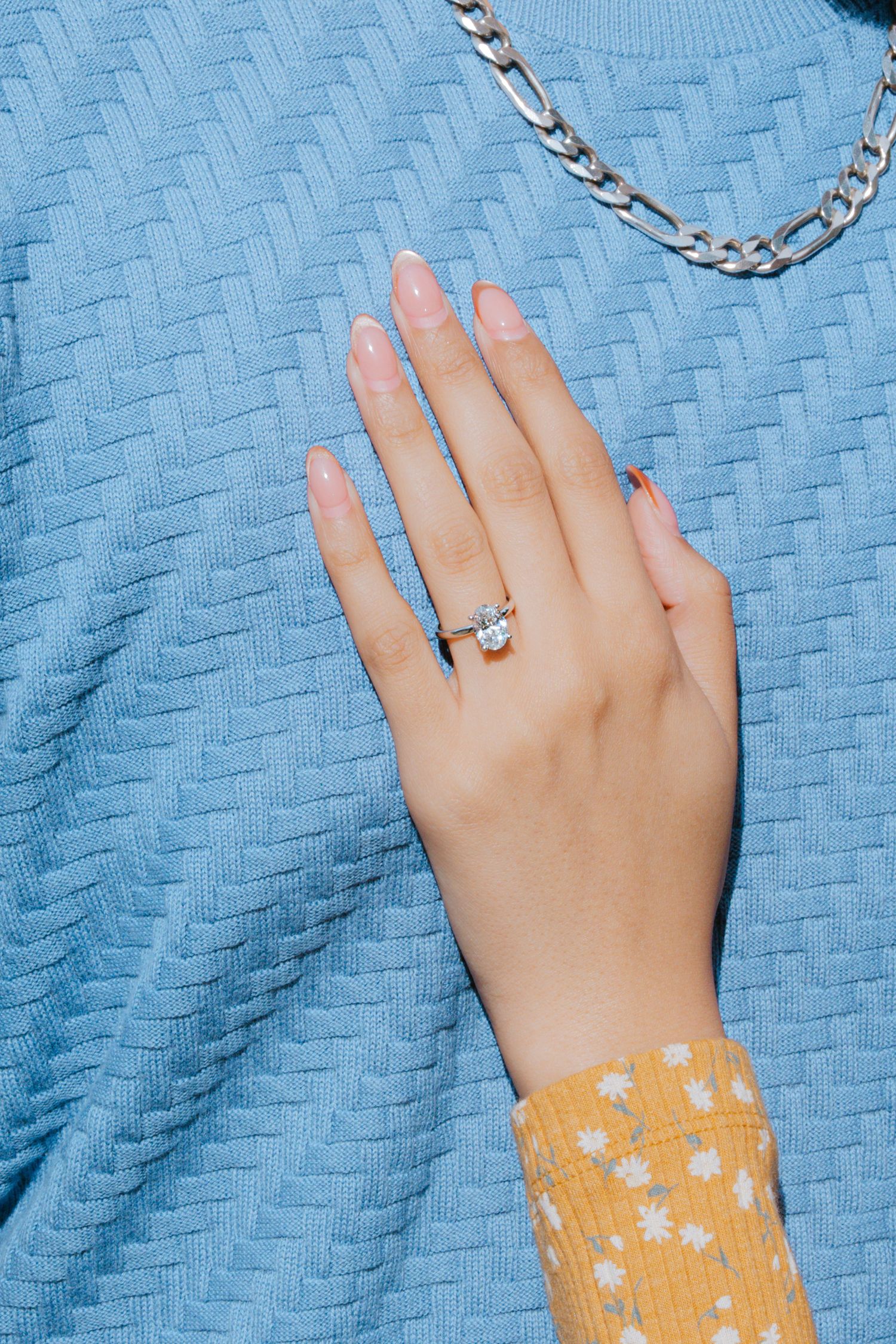 Hand wearing a silver chain bracelet and ring against a blue textured fabric background with a yellow floral sleeve.