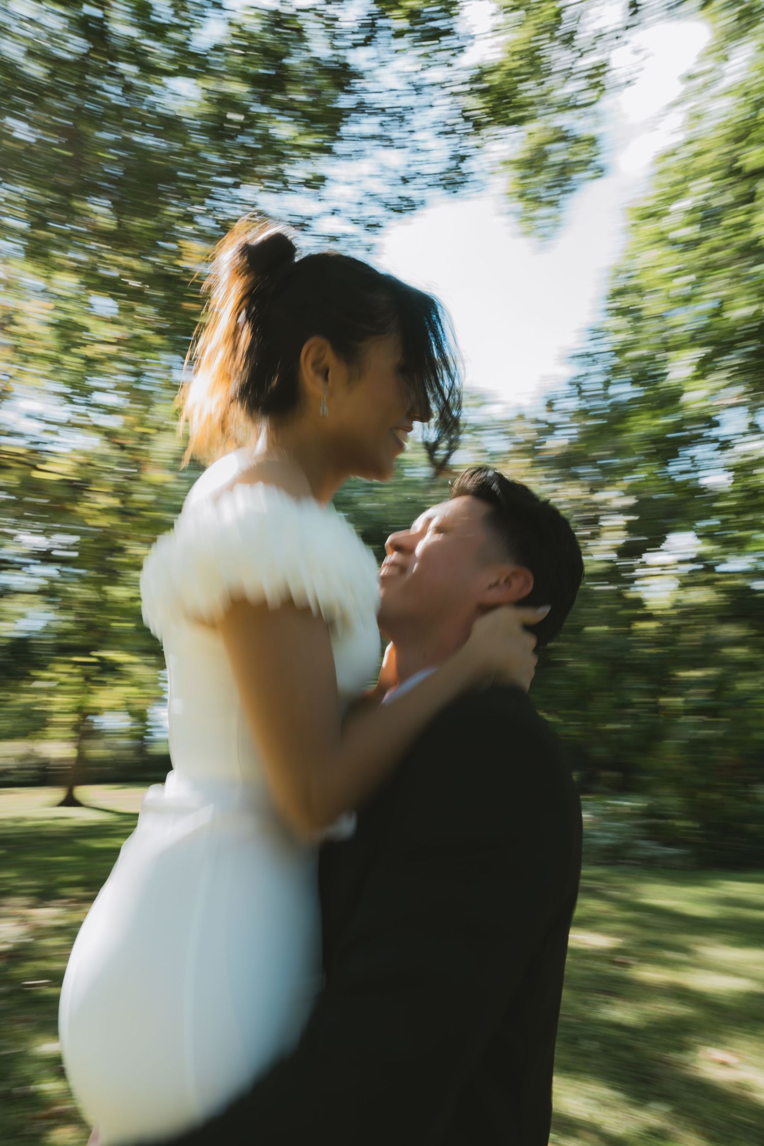 A couple in wedding attire embraces romantically outdoors, surrounded by lush green trees with a dreamy motion blur effect.