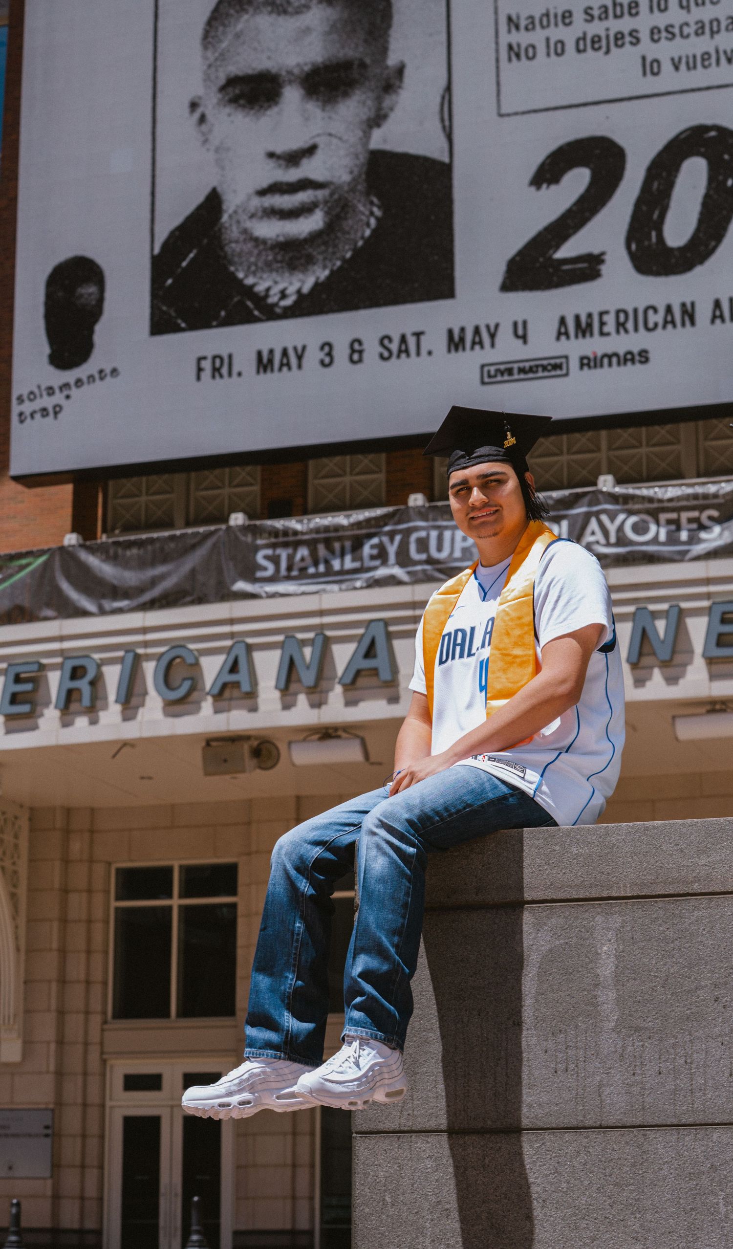 Young man in Dallas Mavericks jersey sits on ledge outside Americana venue with Stanley Cup Playoffs banner.