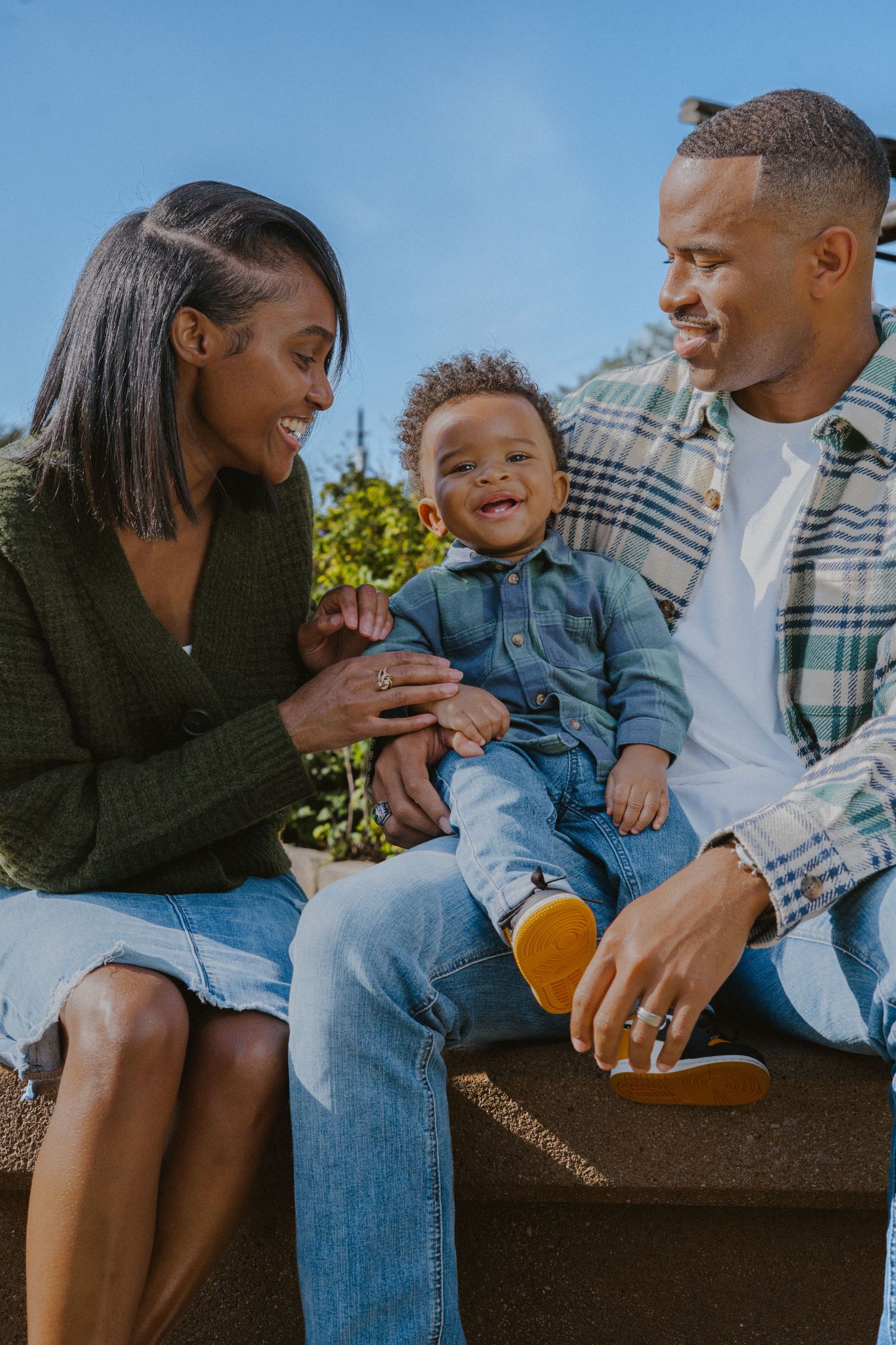 A smiling Black family sits outdoors with their happy toddler boy between them, enjoying a sunny day together.