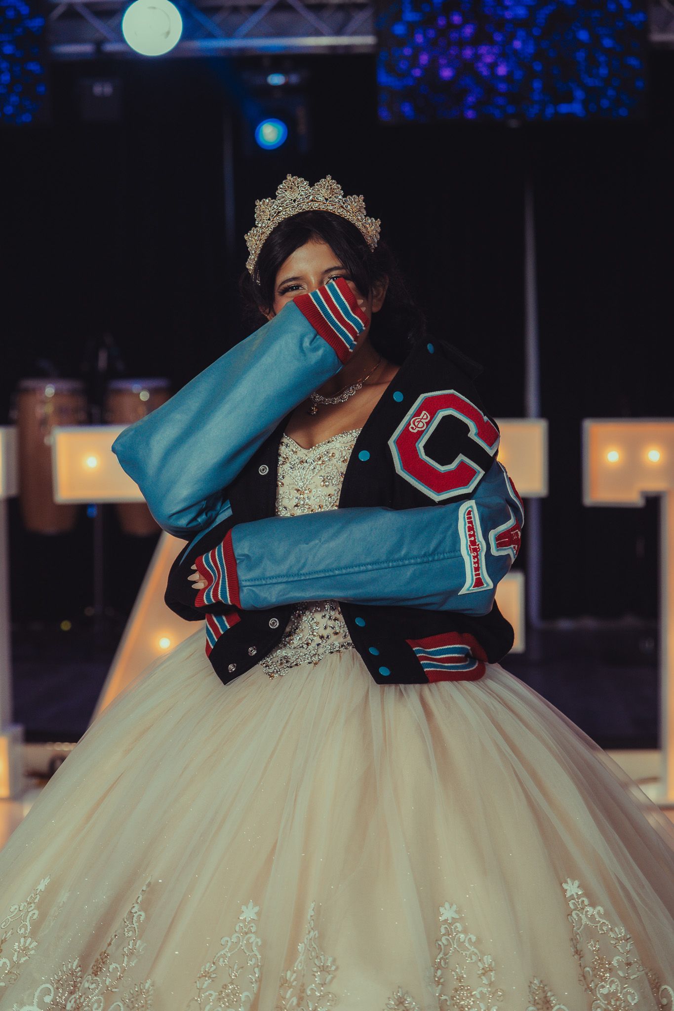 Young woman in quinceañera gown, varsity jacket, and tiara holds blue gift at elegant celebration.