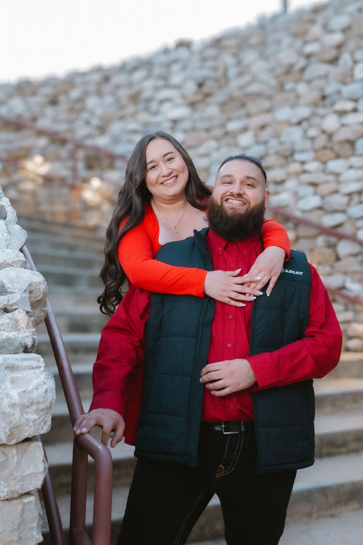 Smiling couple in red outfits embracing on stone steps, woman hugging man with beard from behind.