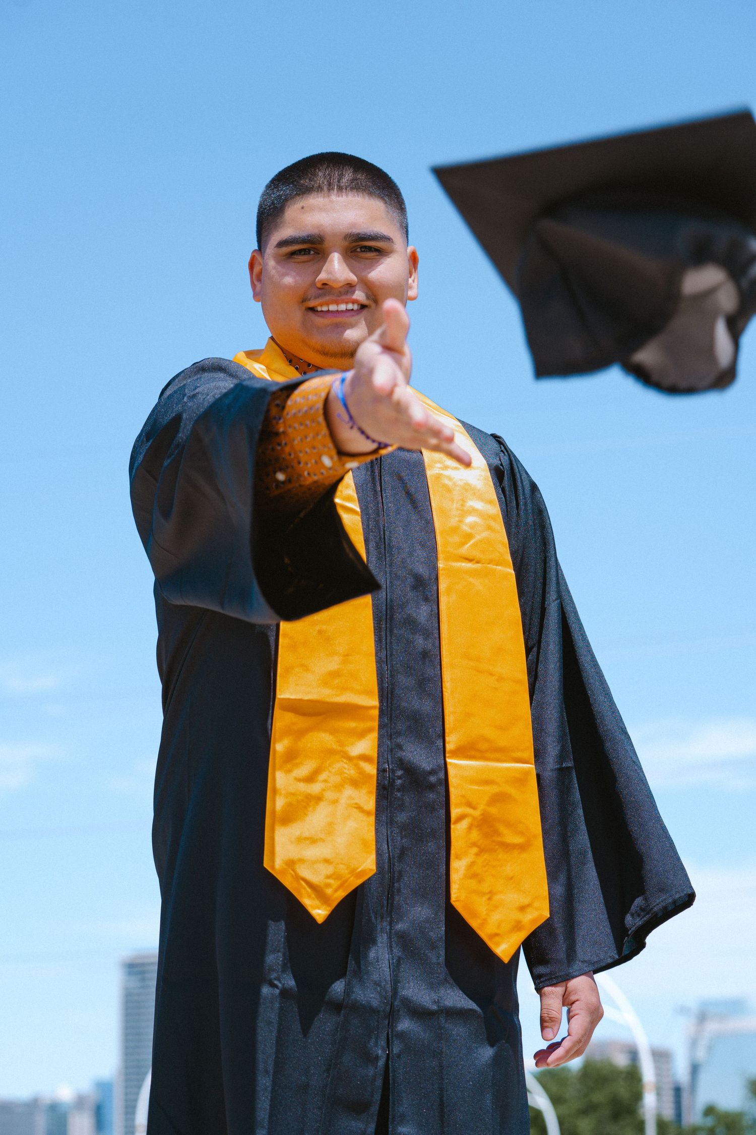 Smiling graduate in black gown and gold stole tosses cap into blue sky, reaching toward camera.