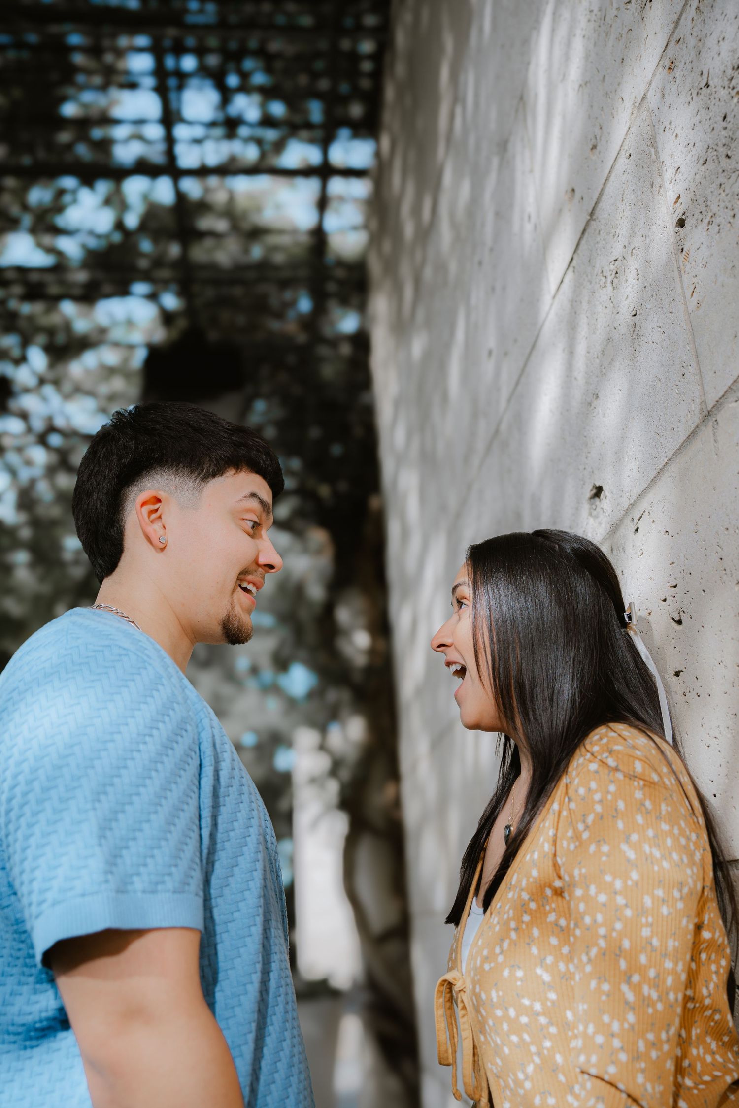 A couple in blue and yellow outfits share intimate glances against a white wall outdoors.