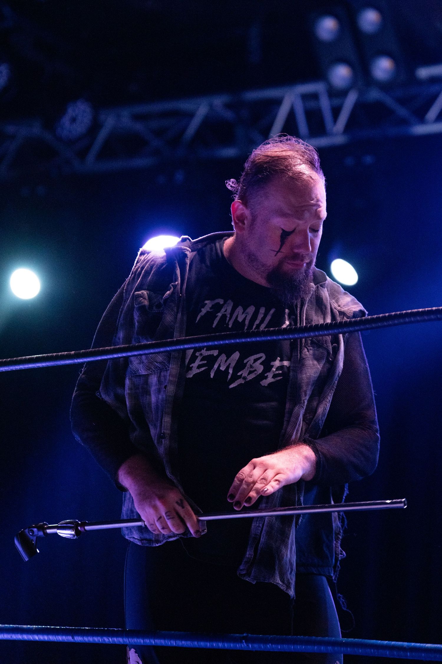 A performer leans against wrestling ring ropes under dramatic blue lighting.