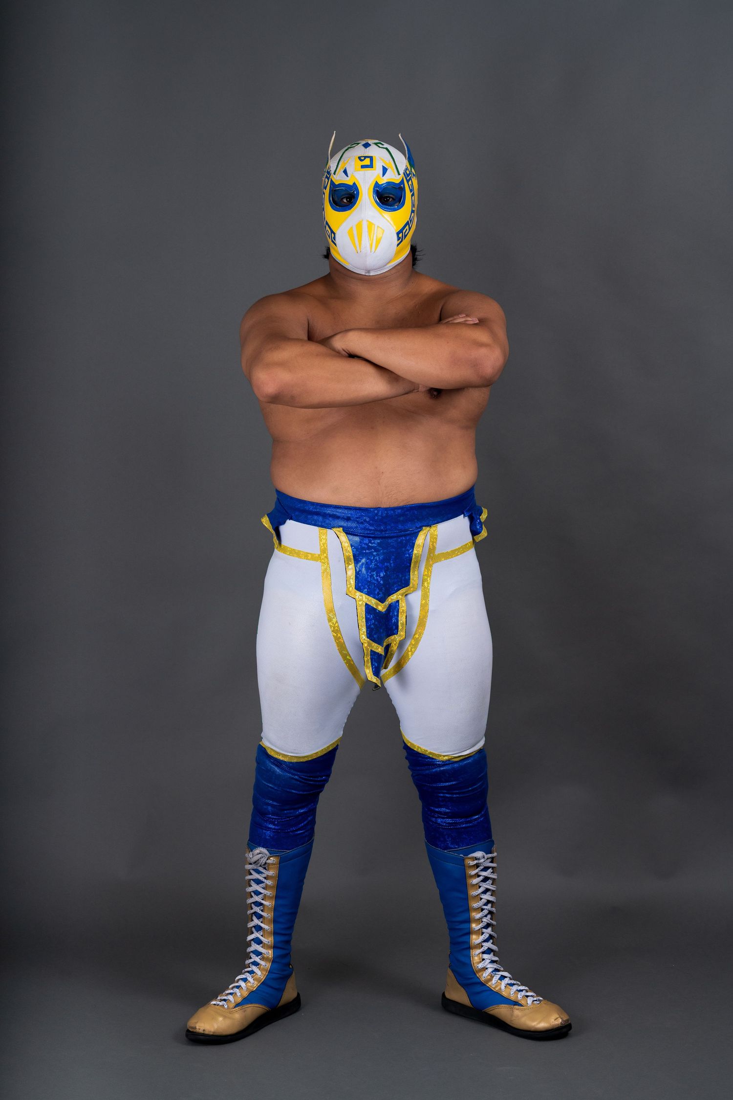 Professional wrestler in blue and white attire with mask striking a power stance against dark background.