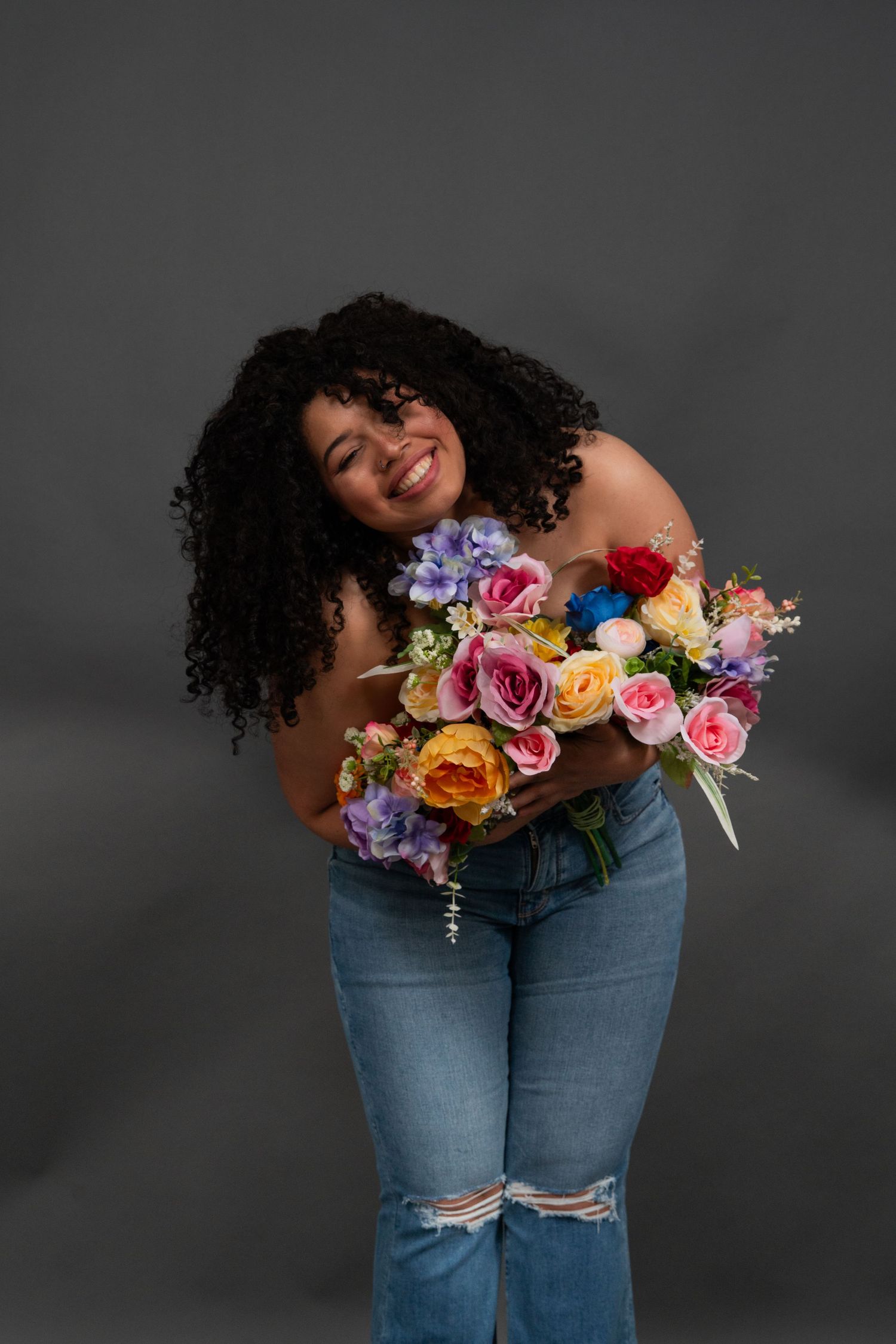 Artistic portrait with colorful floral bouquet and denim jeans against grey backdrop.