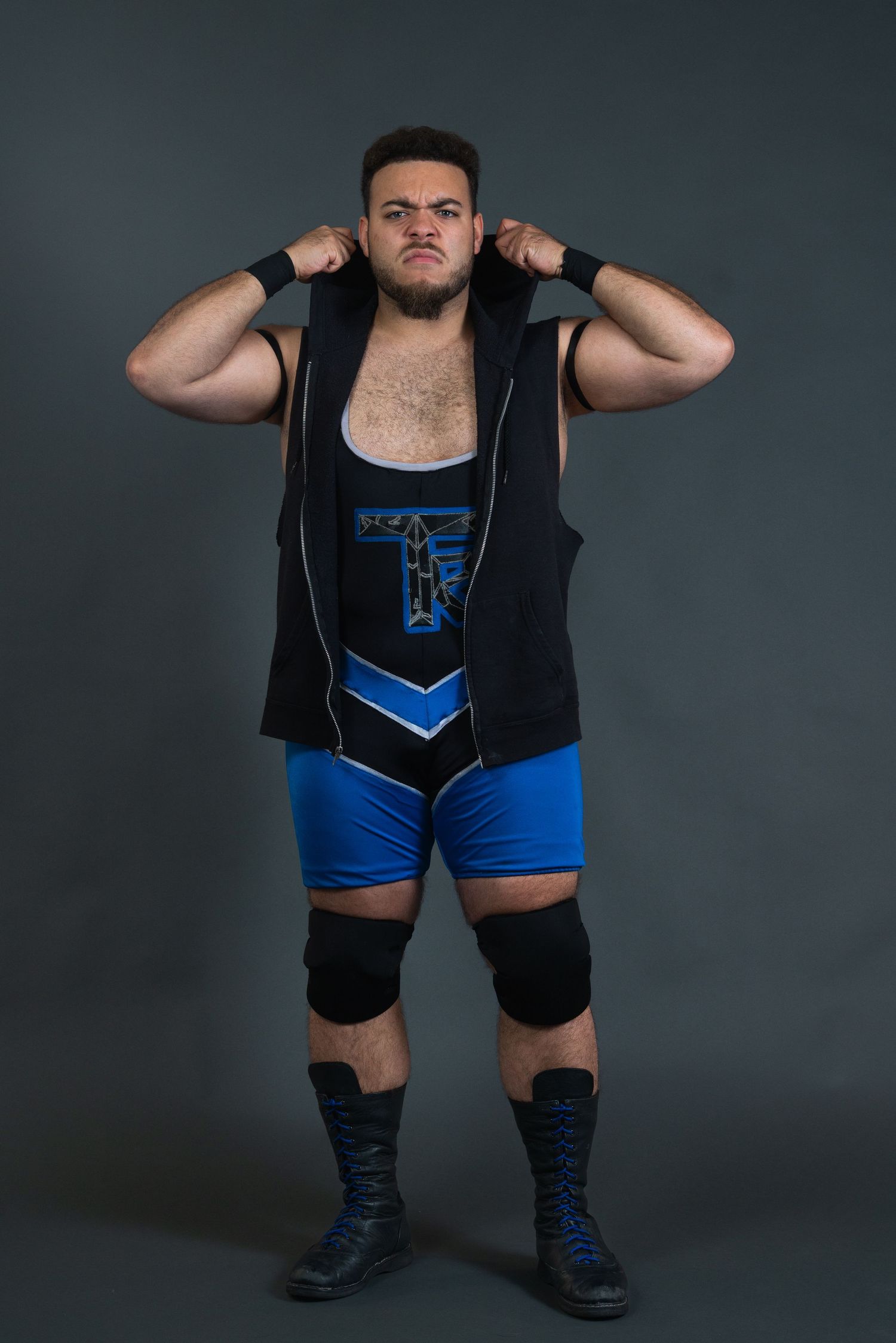 Wrestler wearing black vest and blue shorts poses in studio with dramatic lighting.