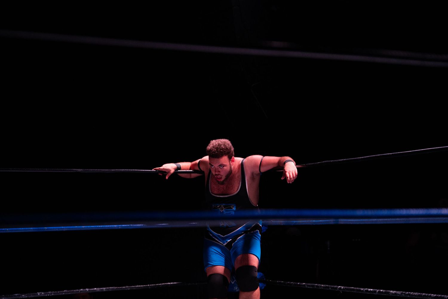 Wrestler executing a move on wrestling ring ropes under dramatic lighting.