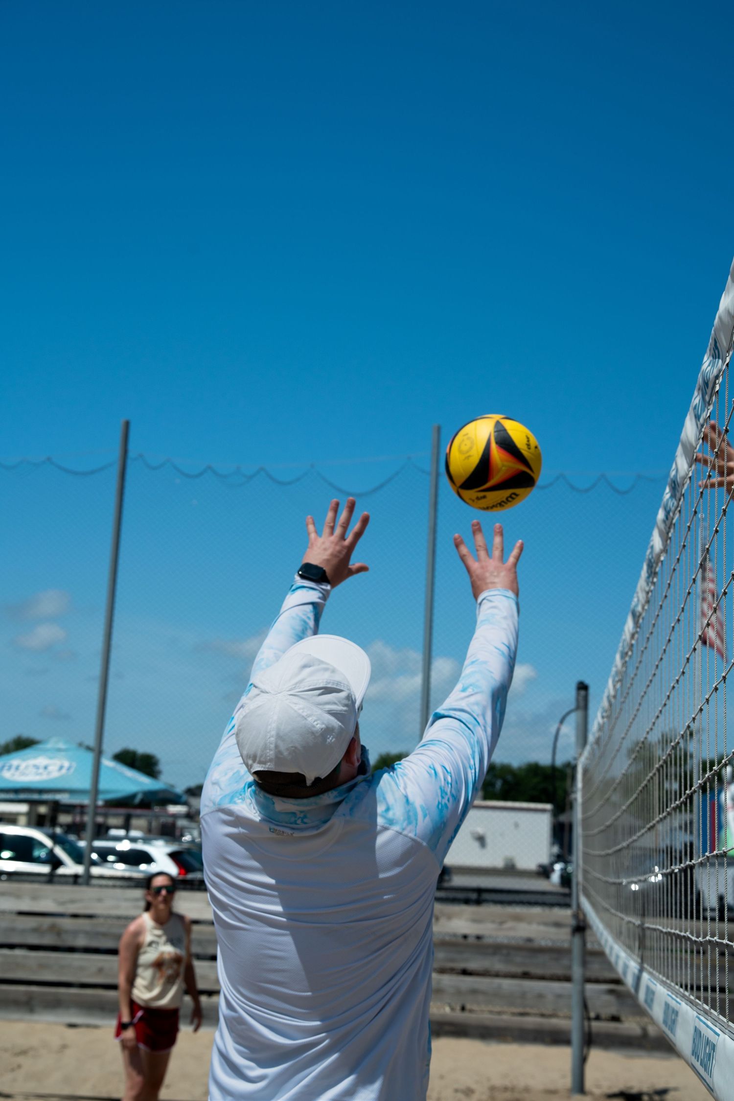 Player in white long sleeves reaching up to hit volleyball against blue sky.