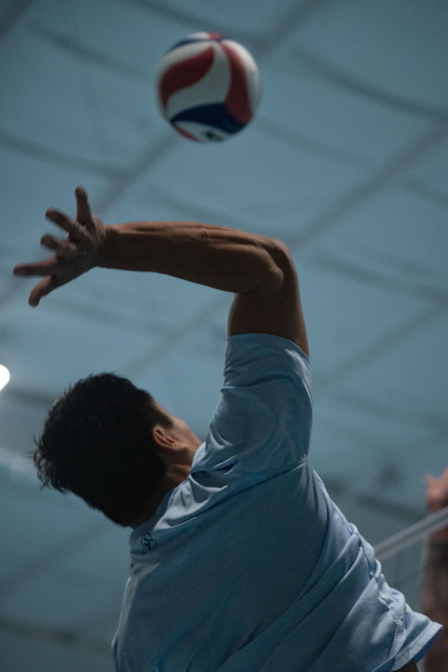 Athletic player jumps to hit a volleyball against a blue indoor ceiling.
