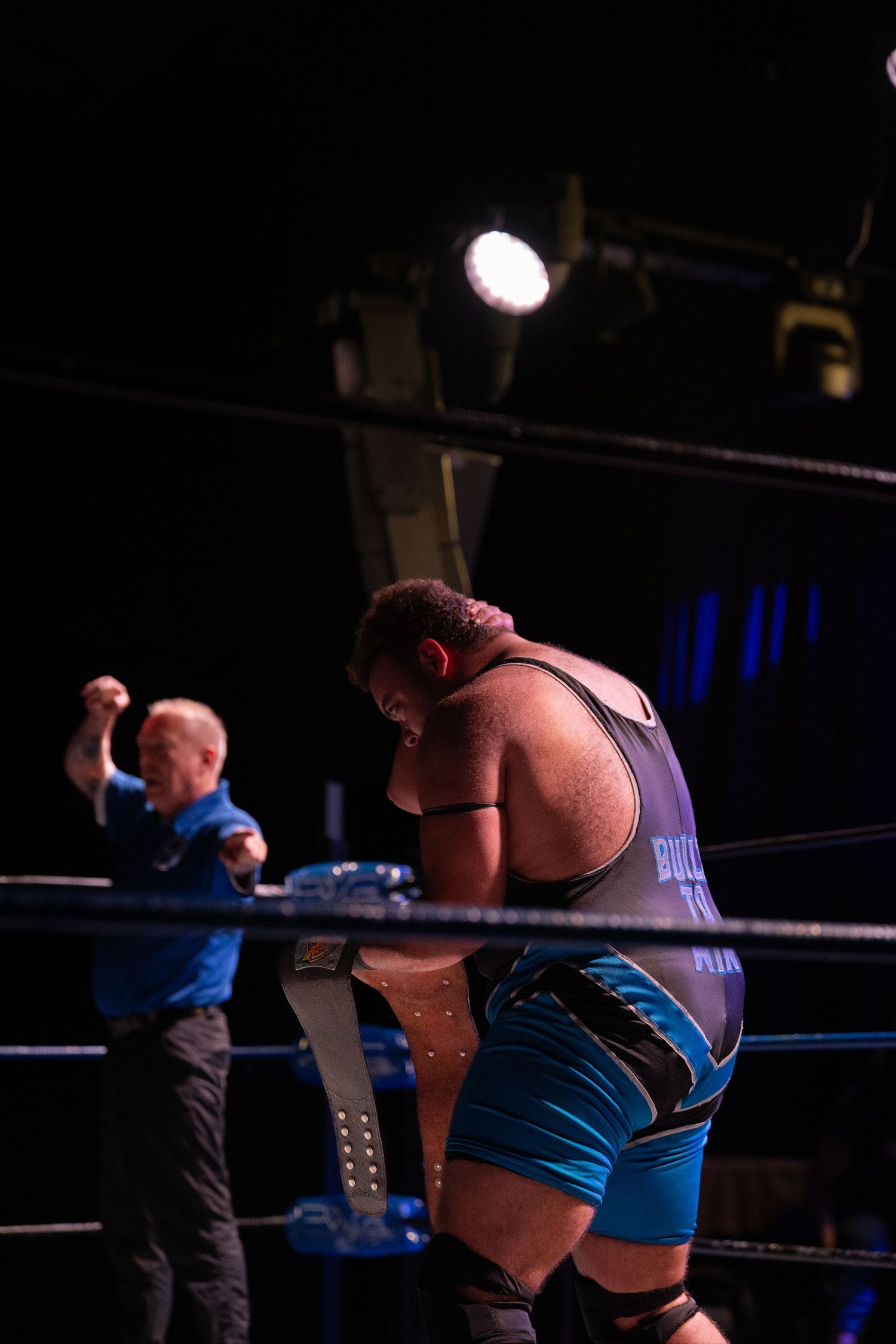 Professional wrestler in blue attire performing in a wrestling ring with referee in background.