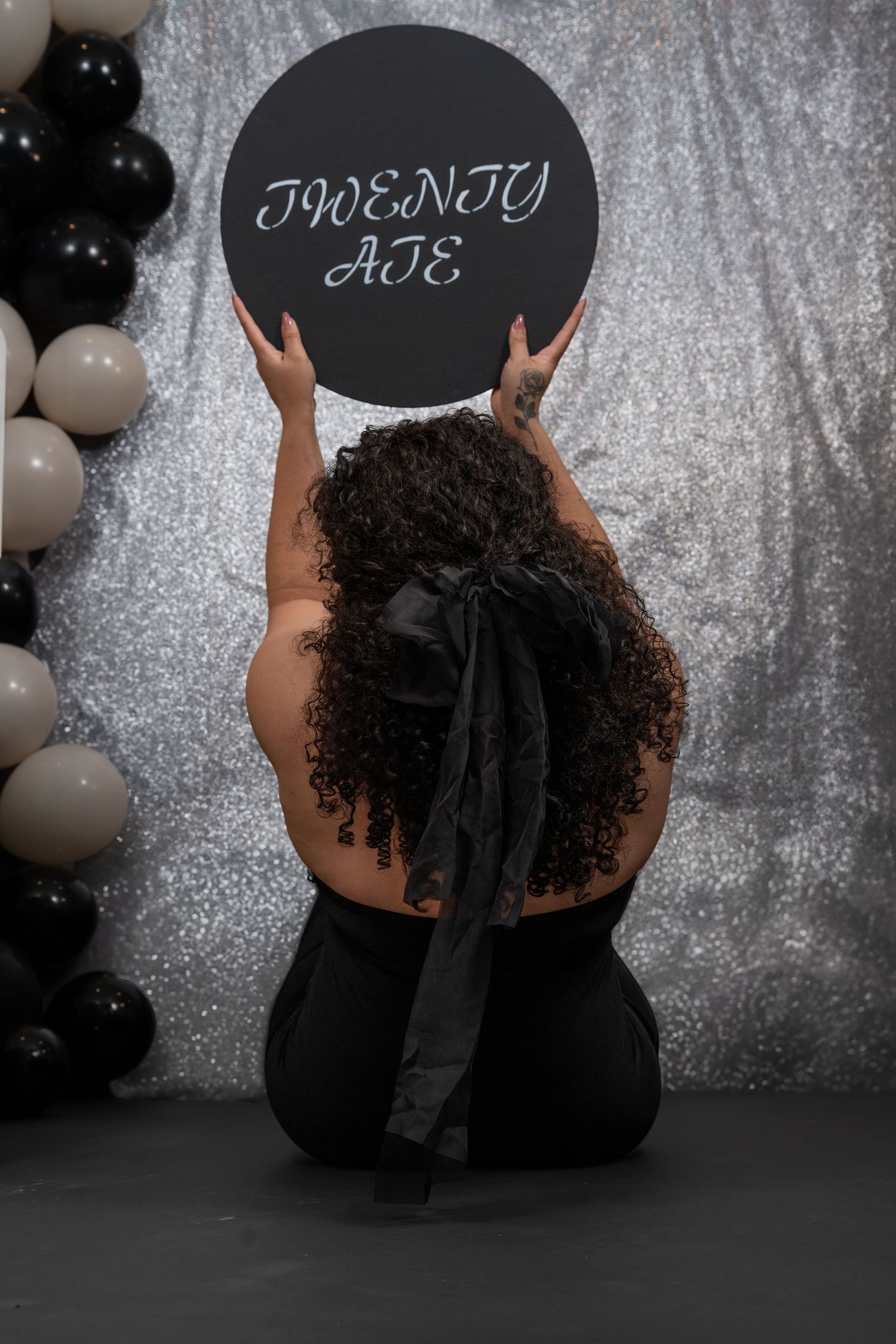 Person in black outfit poses with decorative signs against sparkly silver backdrop with balloons.