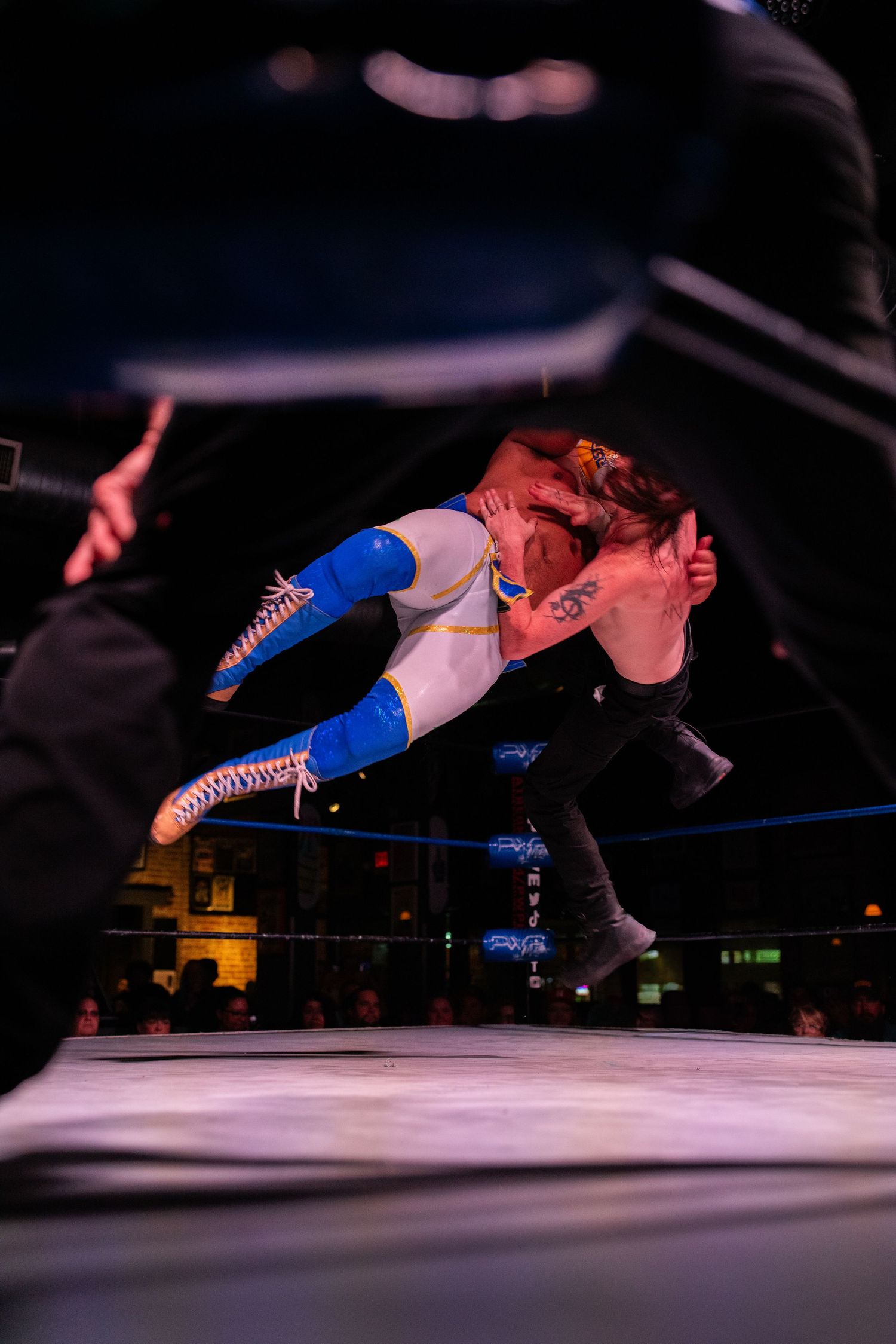 Professional wrestler executes a dramatic aerial maneuver during a match in a dimly lit arena.