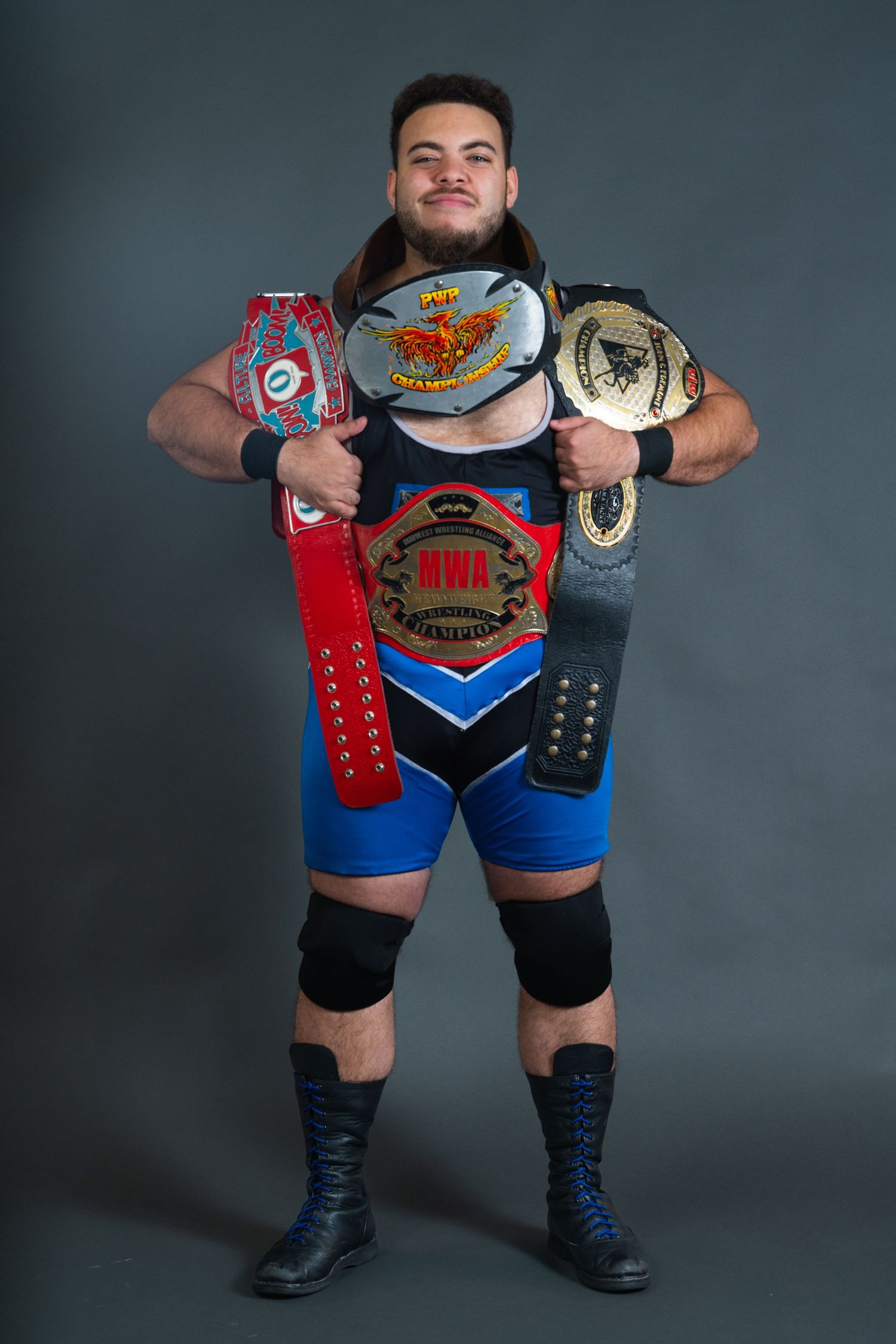 Wrestler displaying championship belts while wearing blue and black wrestling attire.