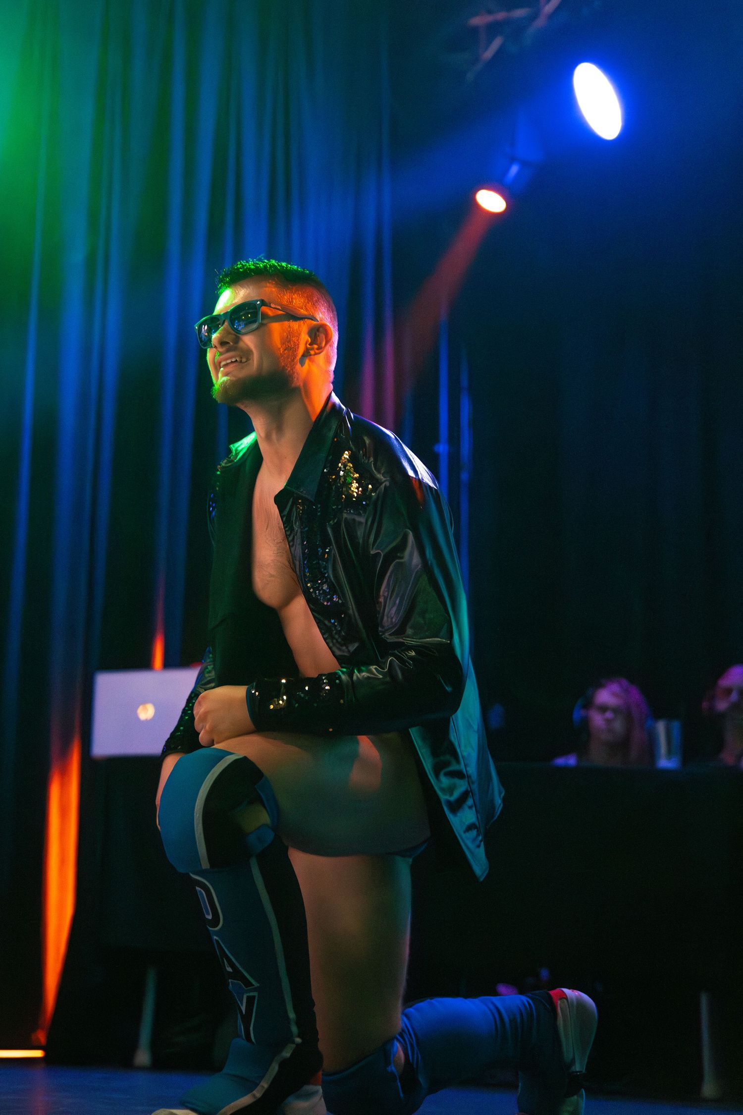 A performer in leather poses dramatically under colorful stage lighting.