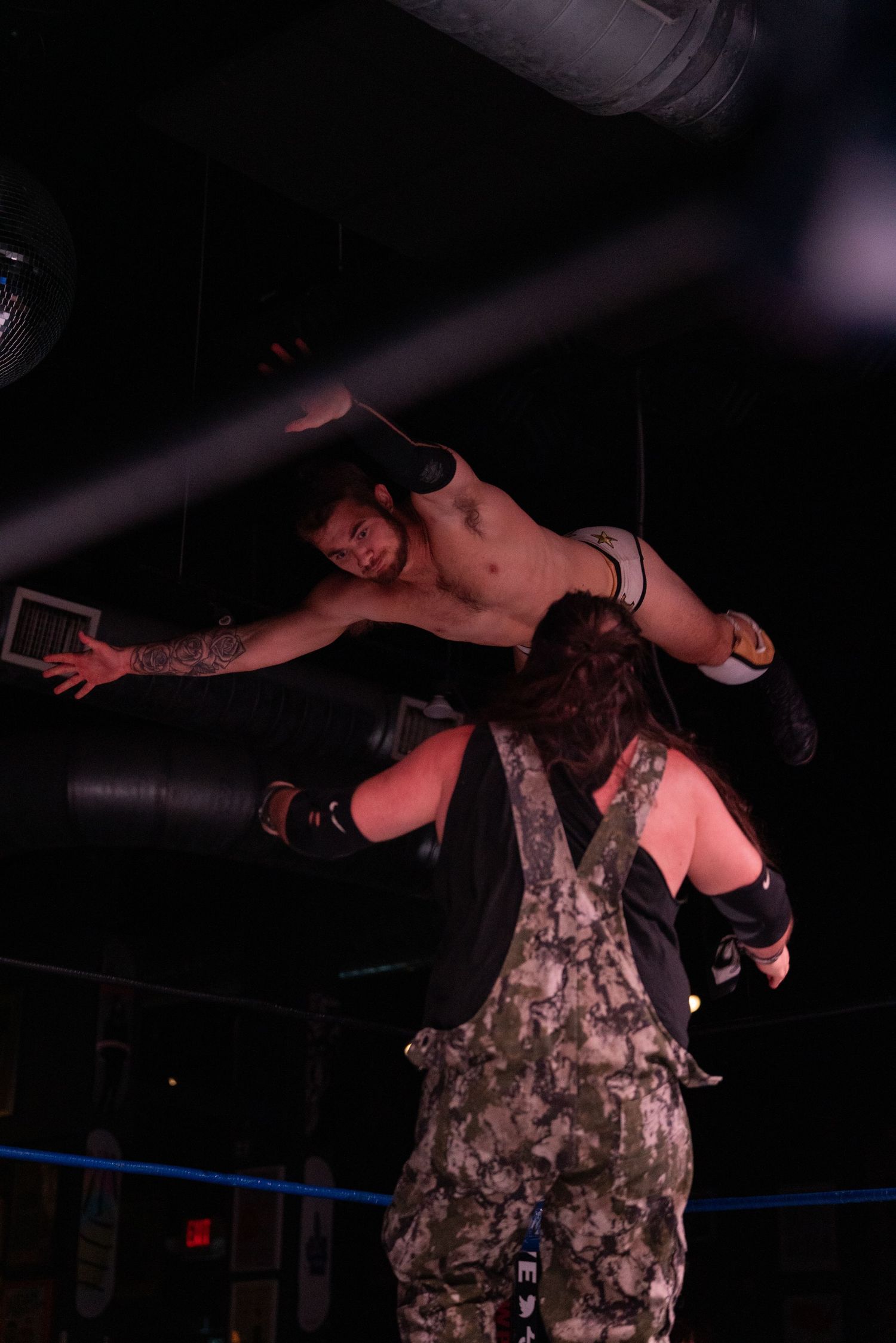Wrestler performs an acrobatic flying move wearing camouflage attire in the ring.