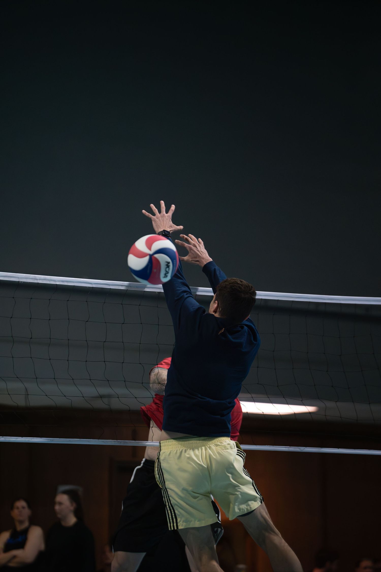 Volleyball players leap high to block a shot at the net during an indoor match.