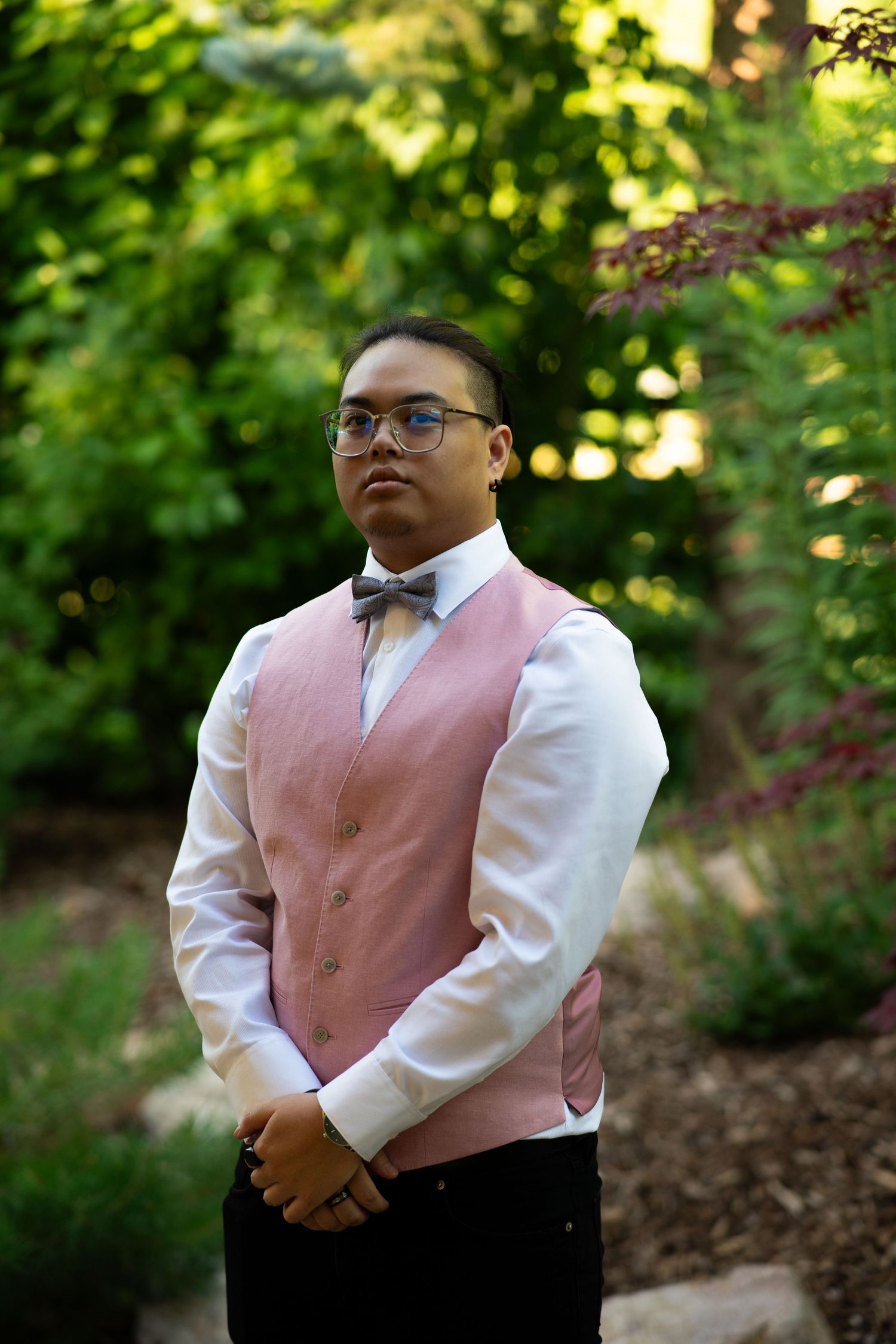 Person wearing pink vest and white dress shirt poses in a lush garden setting.