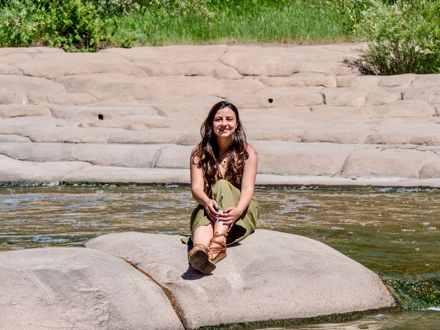 A person sits on a large rock formation by a calm river on a sunny day, with rocky cliffs in the background.