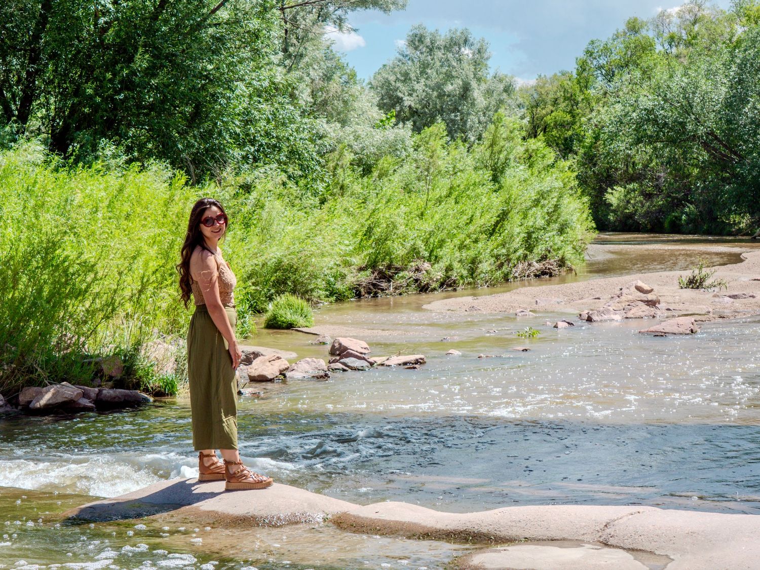Someone walking along the sunny shore of a river with lush green trees and vegetation in the background.