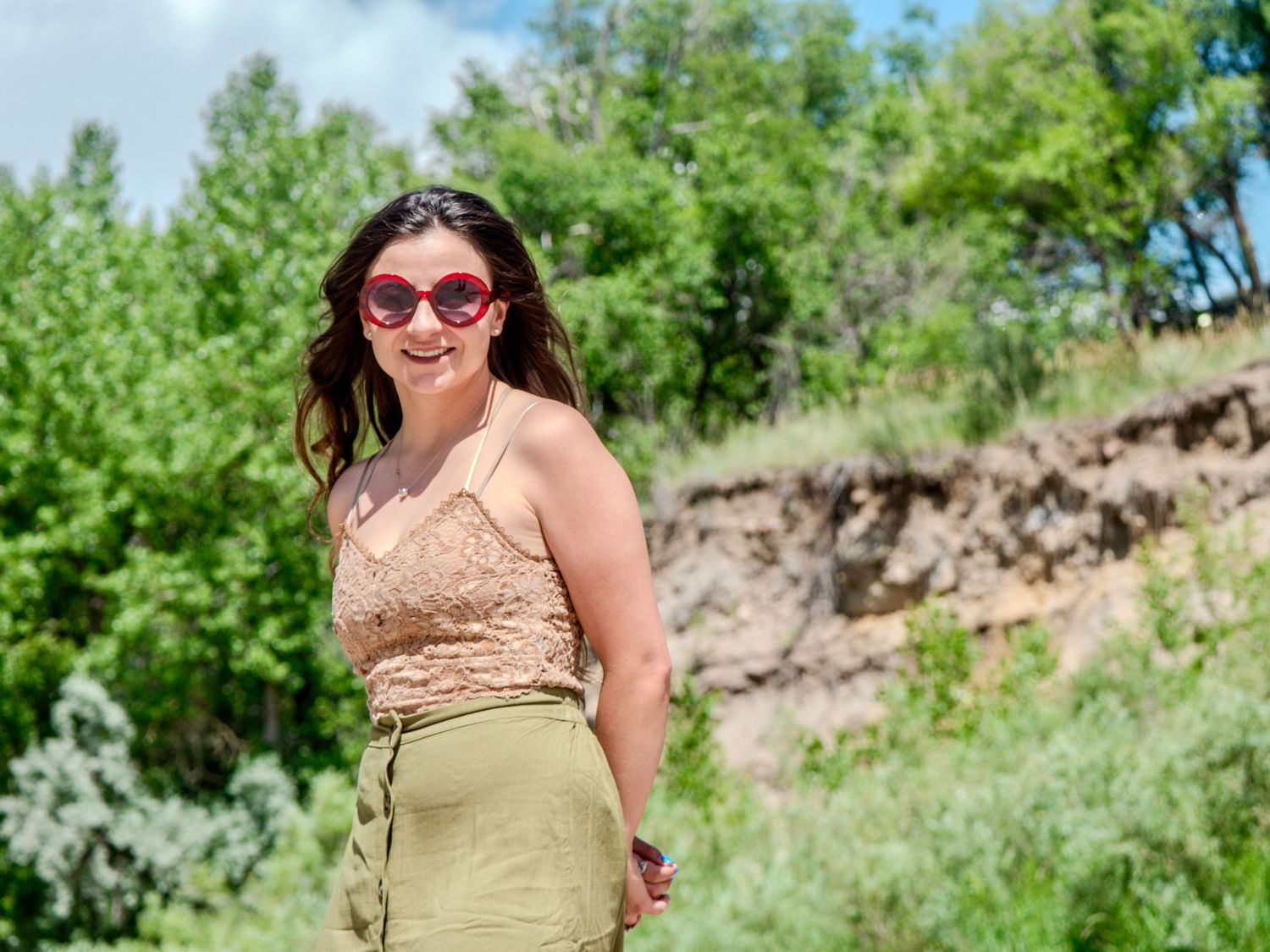 Someone in oversized sunglasses and summer outfit smiles while standing against lush green foliage on a sunny day.