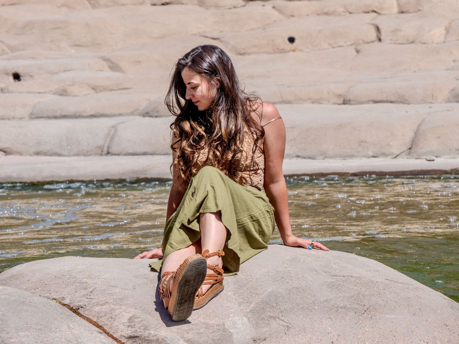 A person in a green skirt and brown top sits on a rock formation near flowing water in a natural setting.