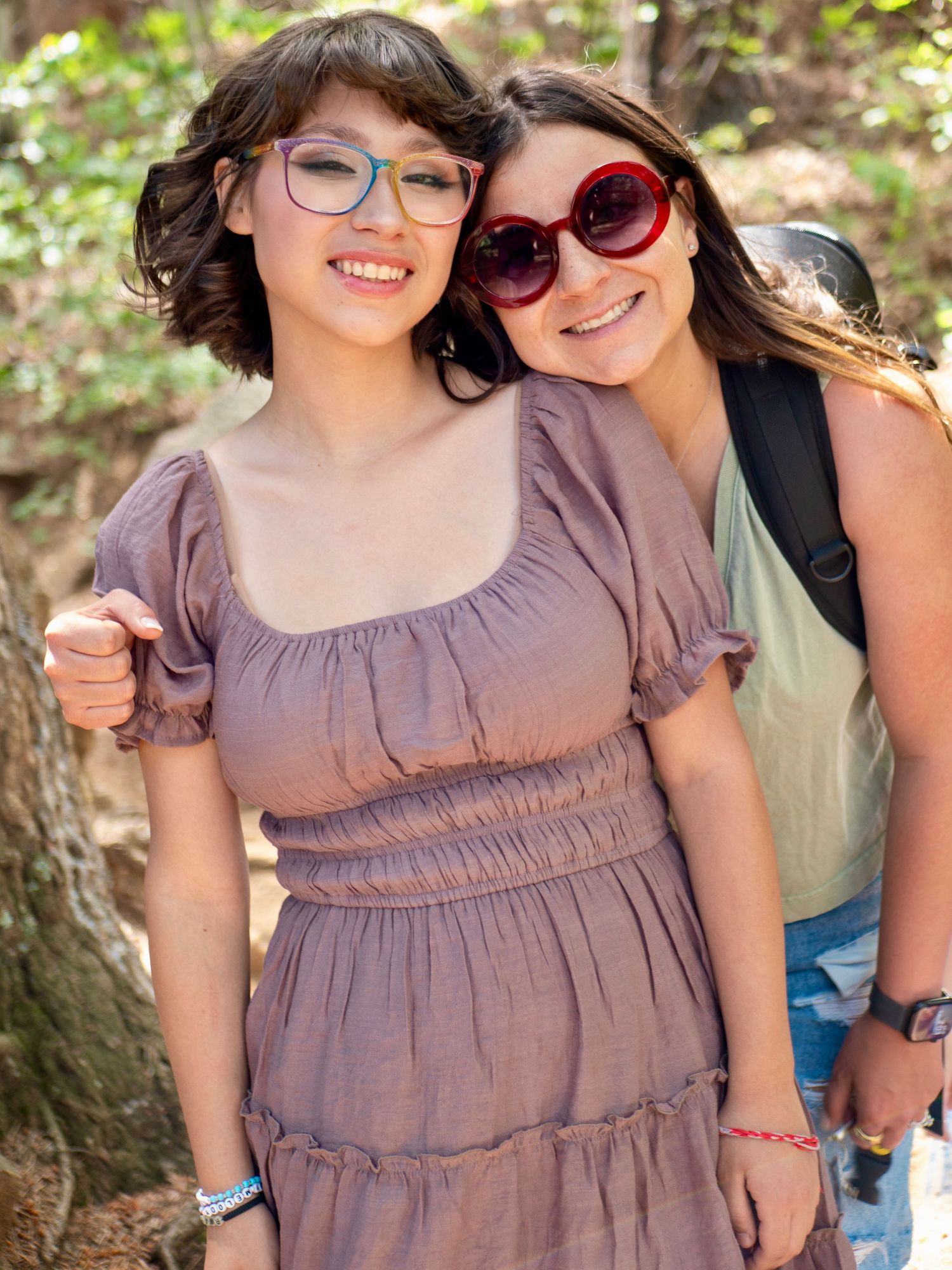 Two people posing together outdoors in casual attire with sunglasses and a mauve dress against a natural background.