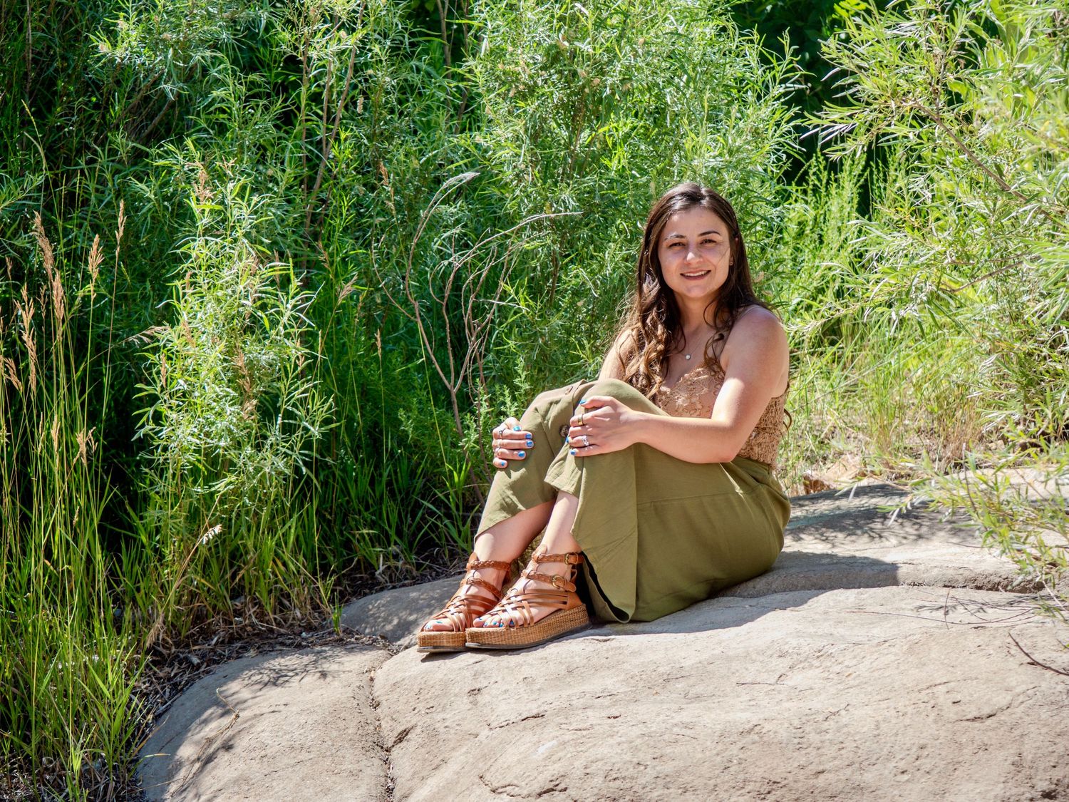 Person sitting on a rocky ledge wearing brown boots and casual clothing in a lush natural outdoor setting.