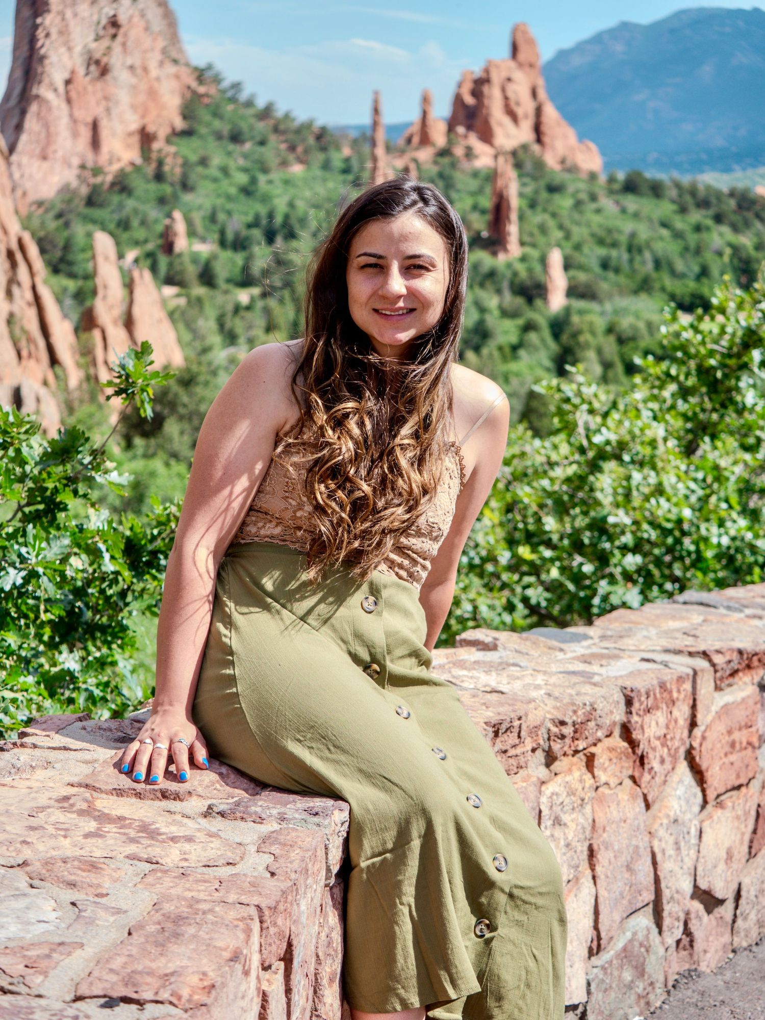A person in a sage green dress poses on red rocks at Garden of the Gods in Colorado with mountains behind.