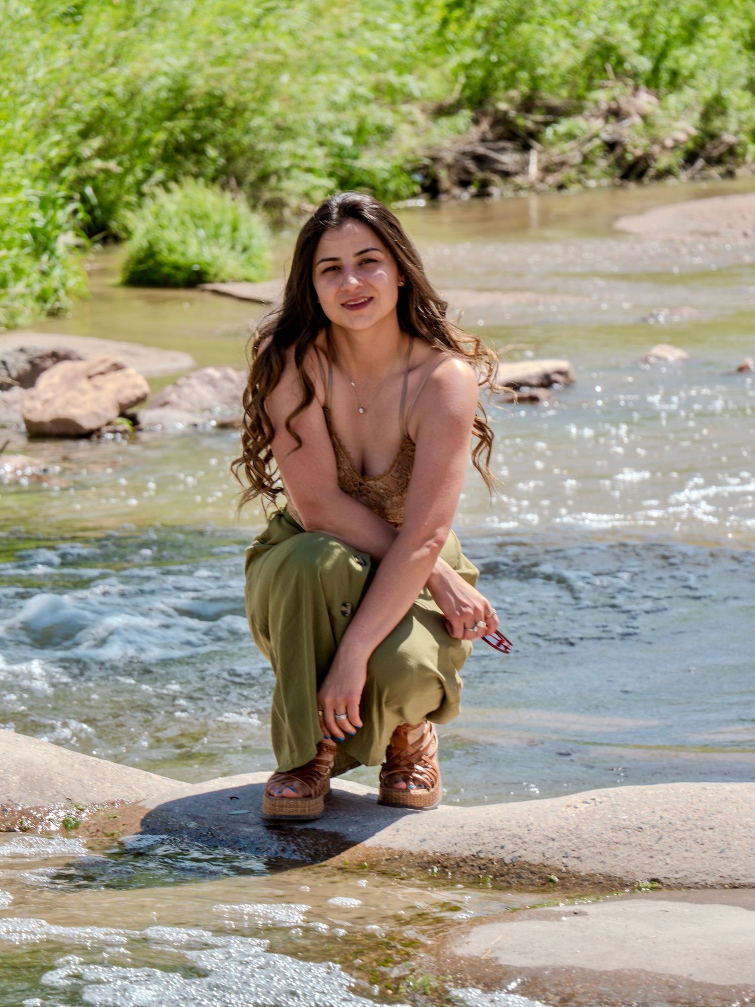 A woman crouches beside a sunlit stream wearing a green skirt and brown boots among rocks and flowing water.