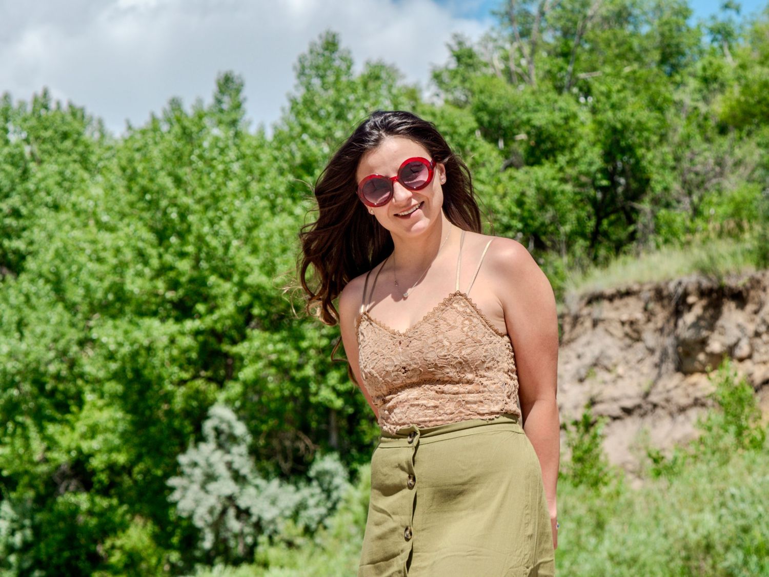 A person in heart-shaped sunglasses and beige top poses outdoors on a sunny day with green trees in the background.
