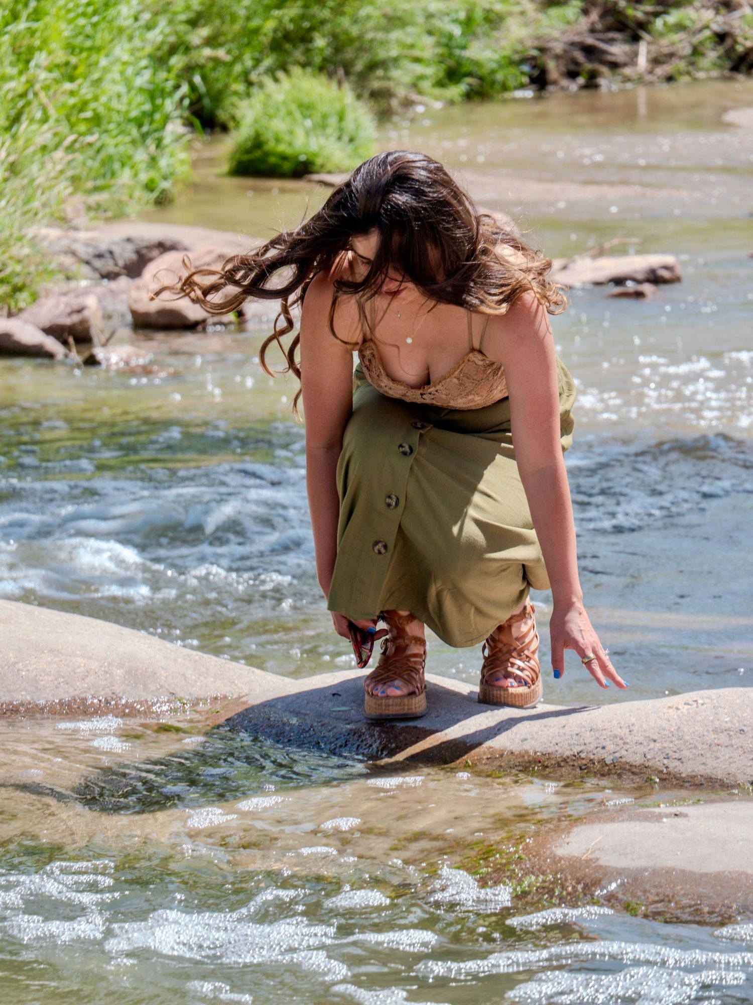 A person in olive clothing crouches by a river stream to interact with flowing water on a sunny day.