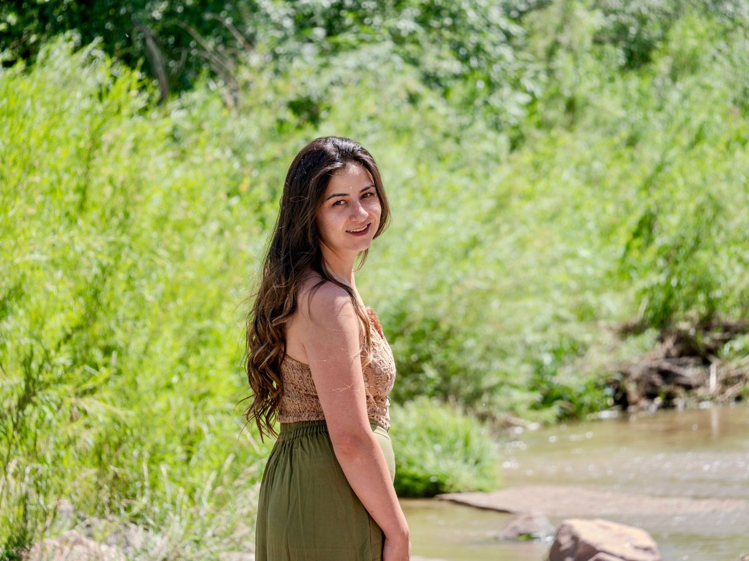 A figure in an olive green dress stands beside a stream surrounded by lush summer greenery in nature.