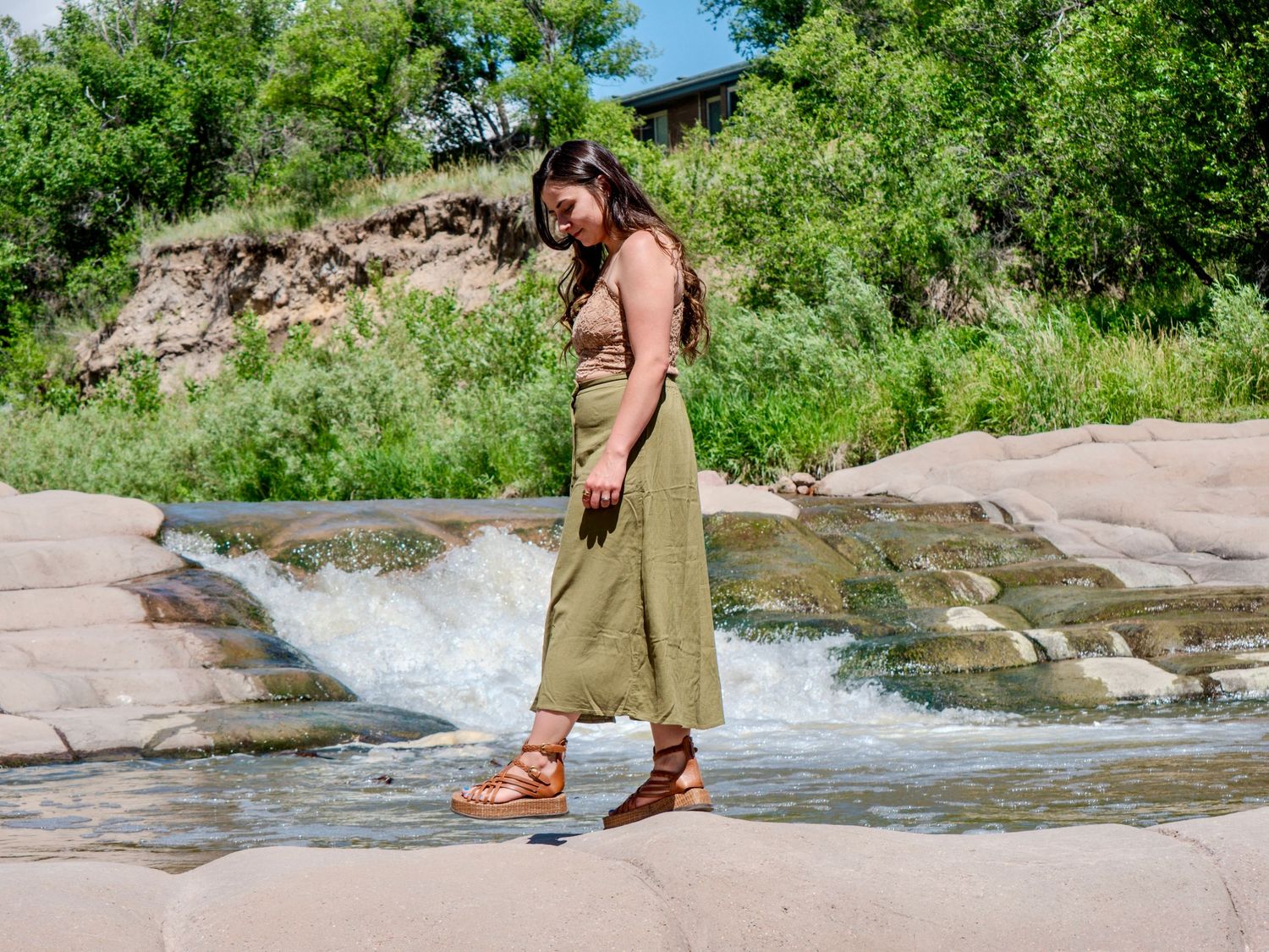 Someone in a long olive dress stands next to flowing water on pink granite rocks surrounded by green foliage.