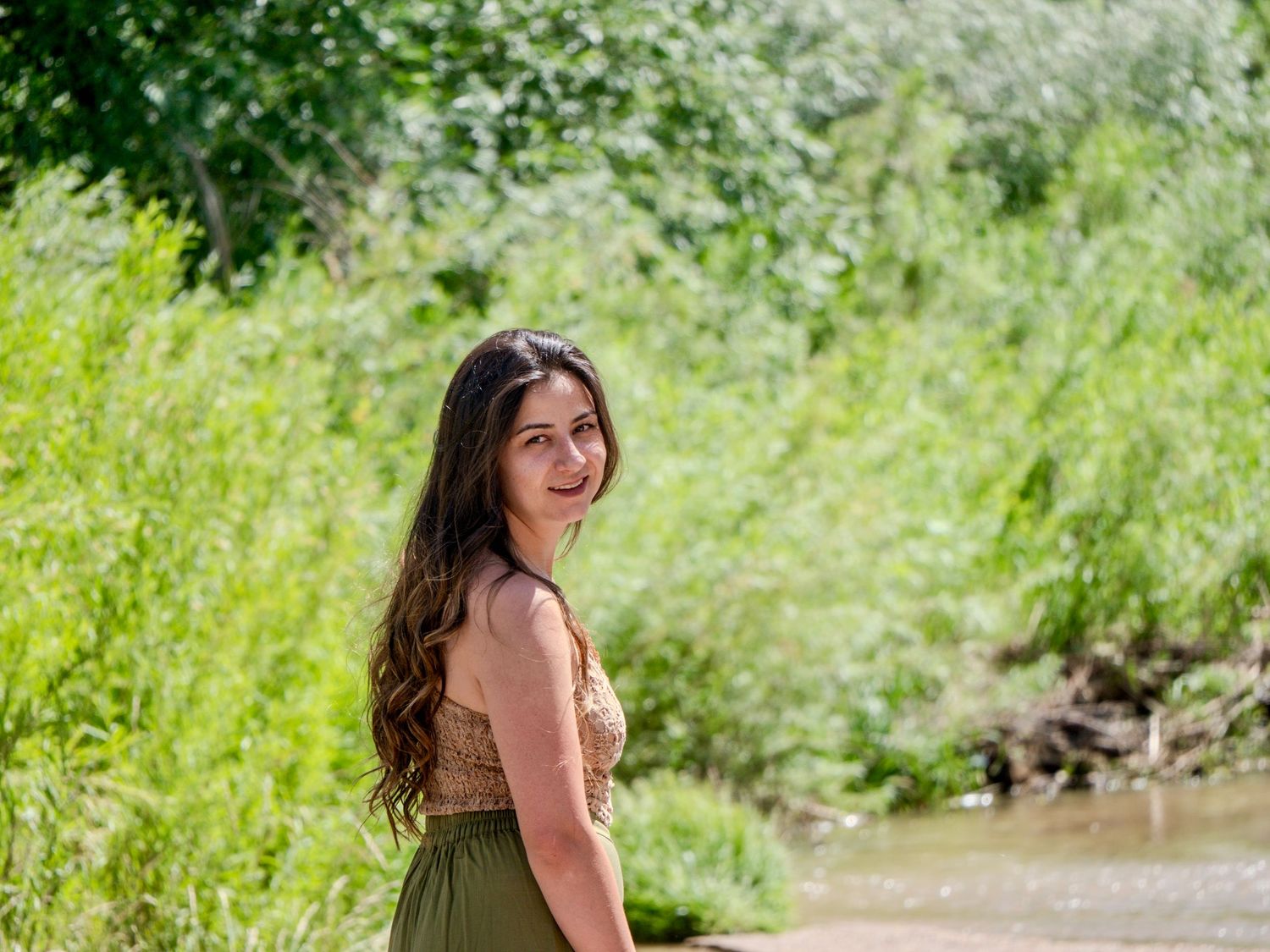 A person in an olive green dress stands on a trail surrounded by lush greenery on a sunny summer day.