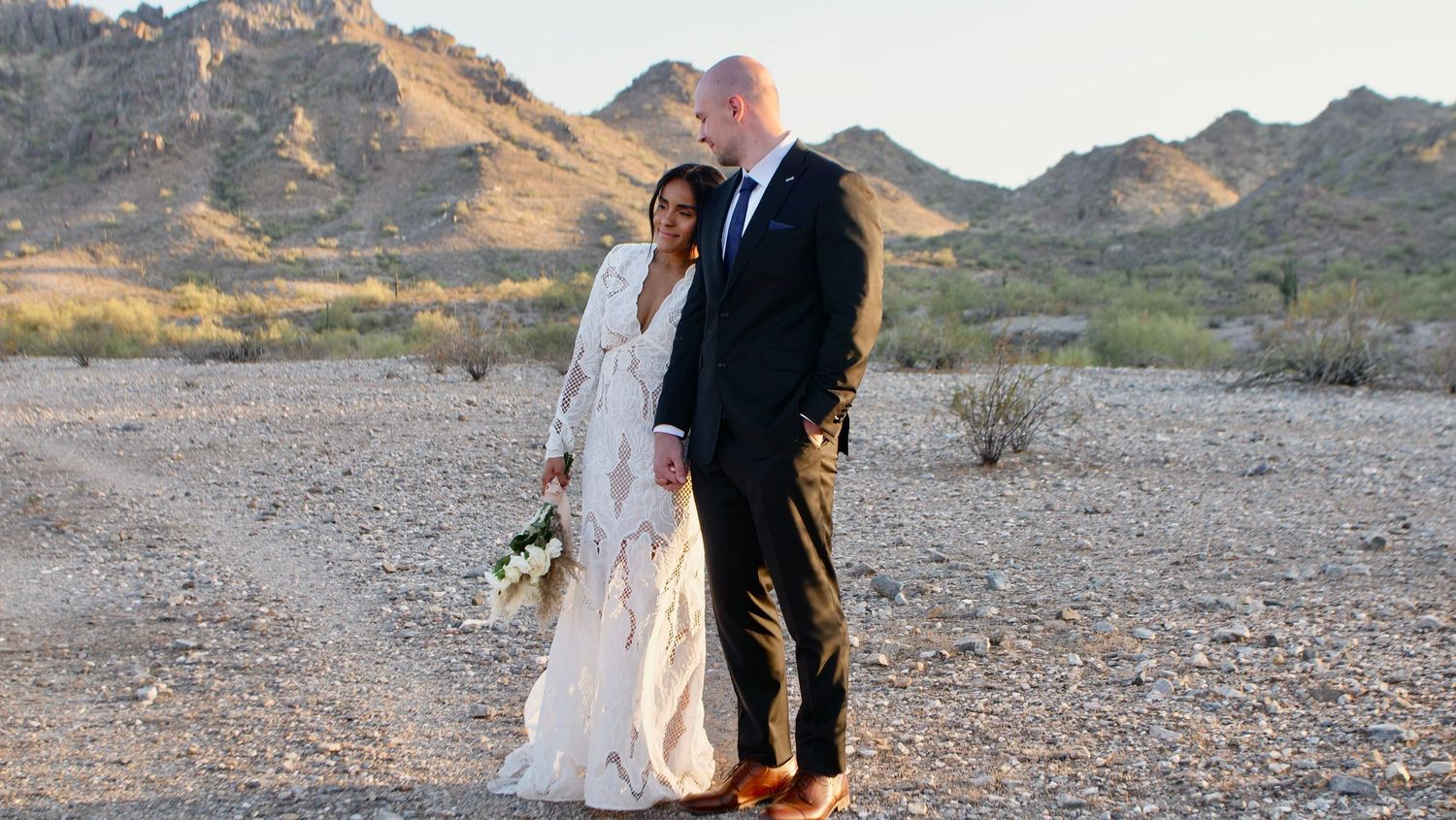 A couple in wedding attire embrace while standing in a desert landscape with rocky mountains in the background at sunset.