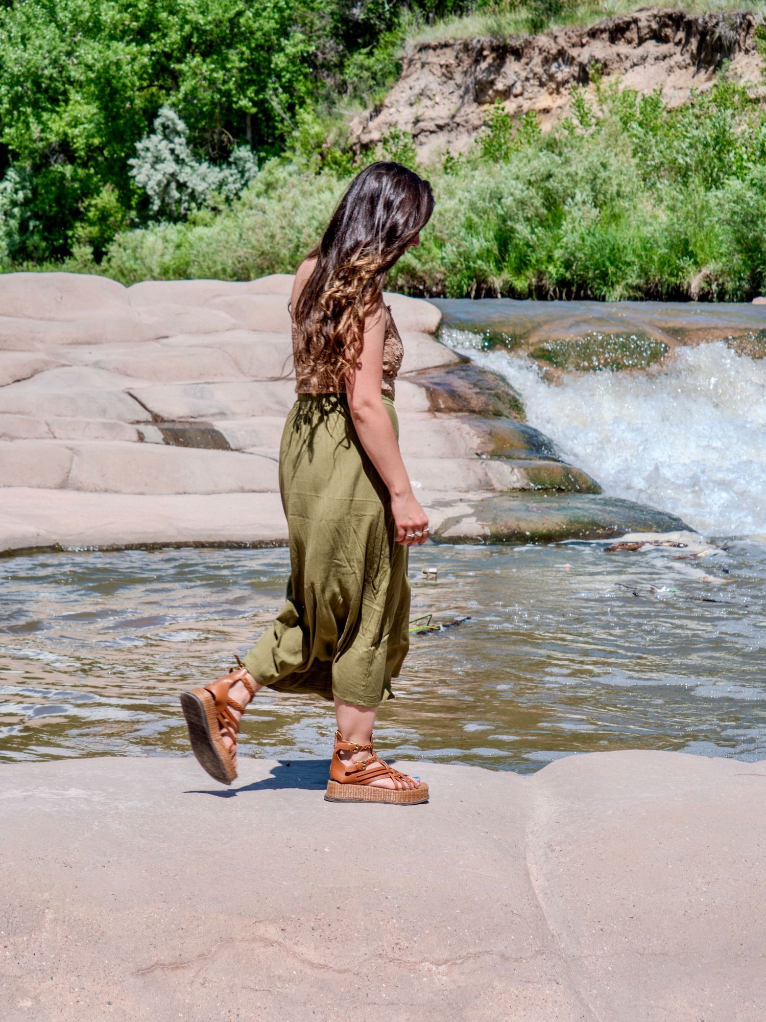 A person in an olive green dress and sandals stands on wet rocks near rushing water on a sunny summer day.